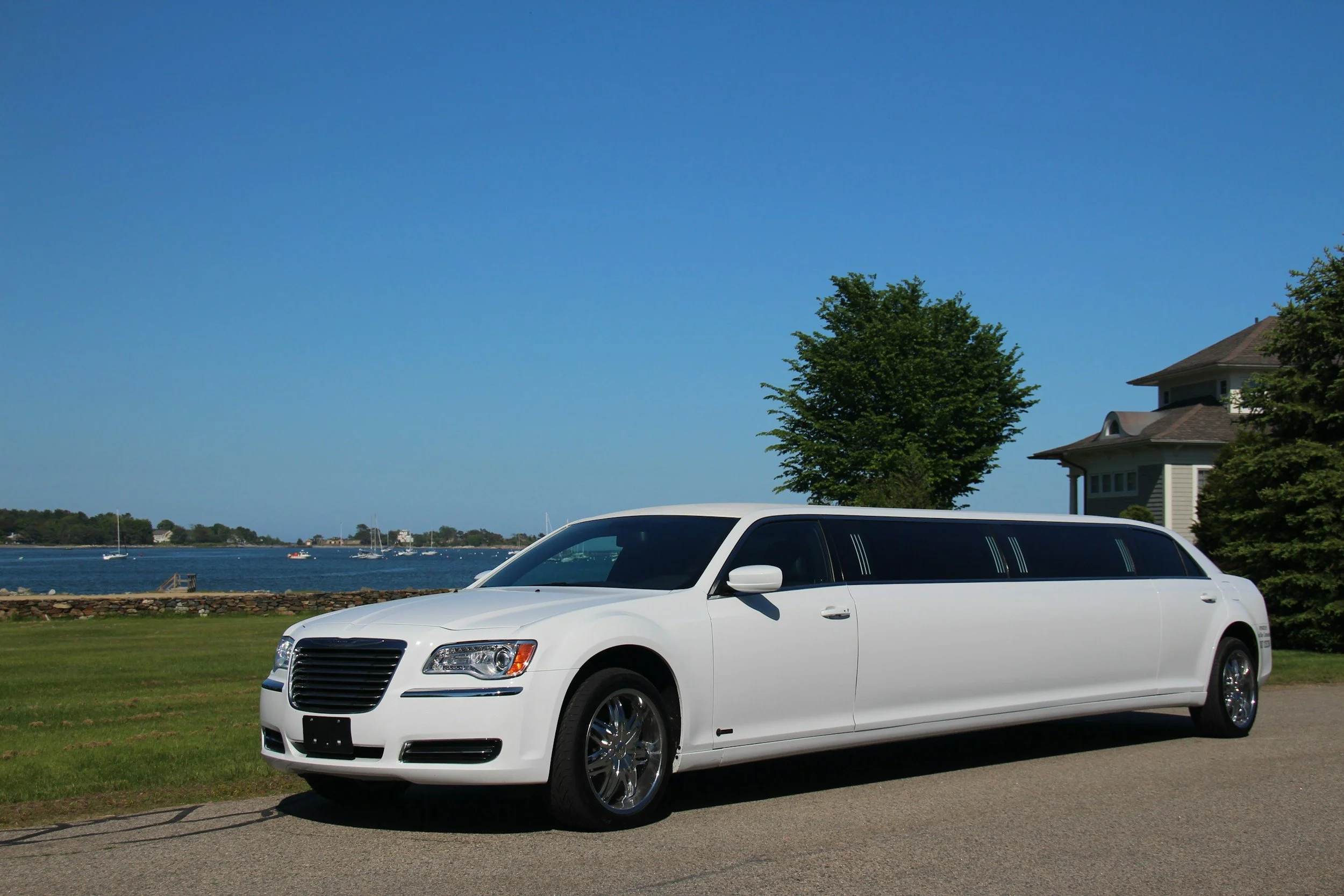 A white limousine parked on a paved area near a grassy field with water in the background. There are sailboats on the water, and a house with a peaked roof and a large tree nearby under a clear blue sky.