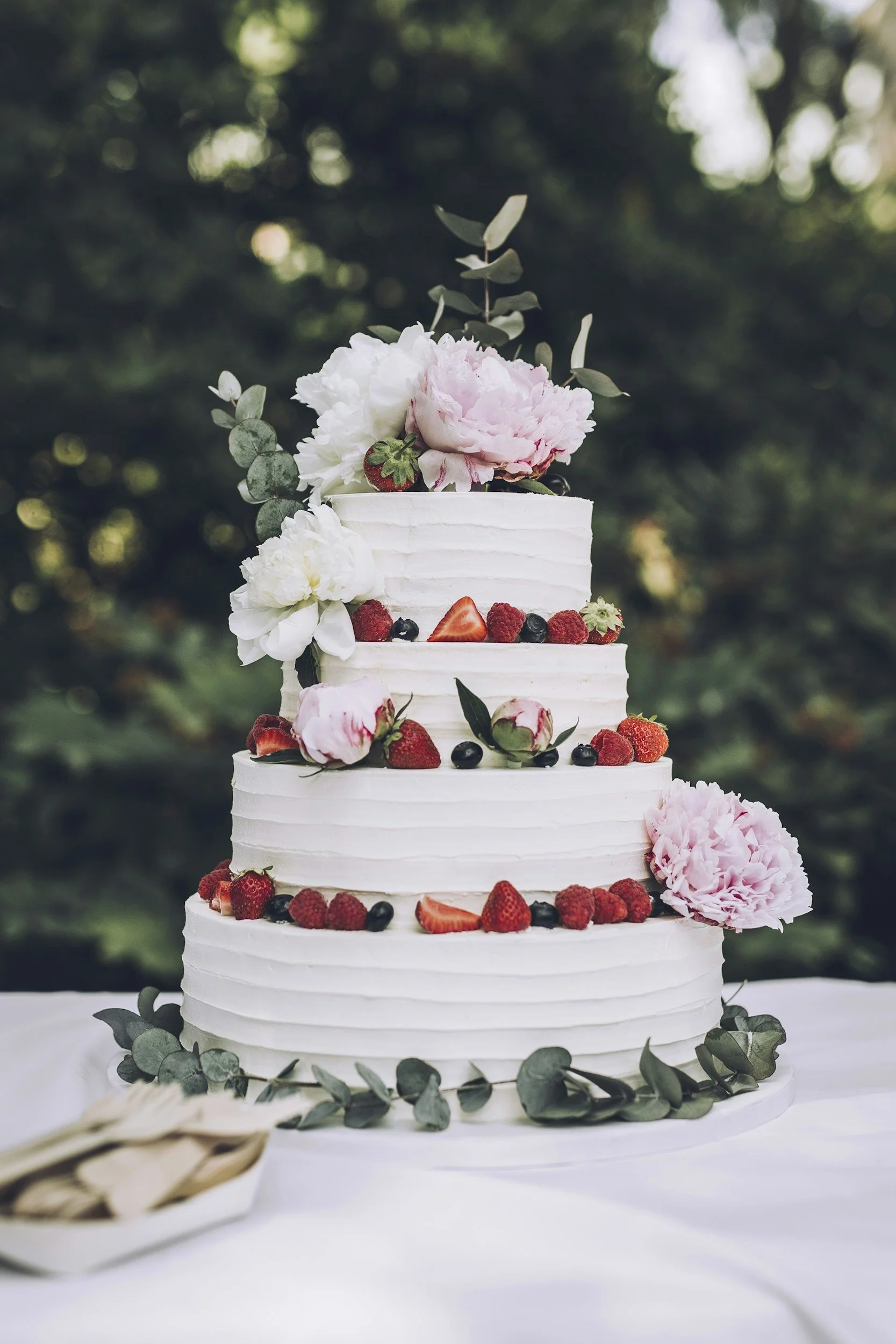 Multi-tiered white wedding cake decorated with fresh strawberries, raspberries, blueberries, white and pink peonies, and lavenders, set outdoors on a white table with a green blurred background.