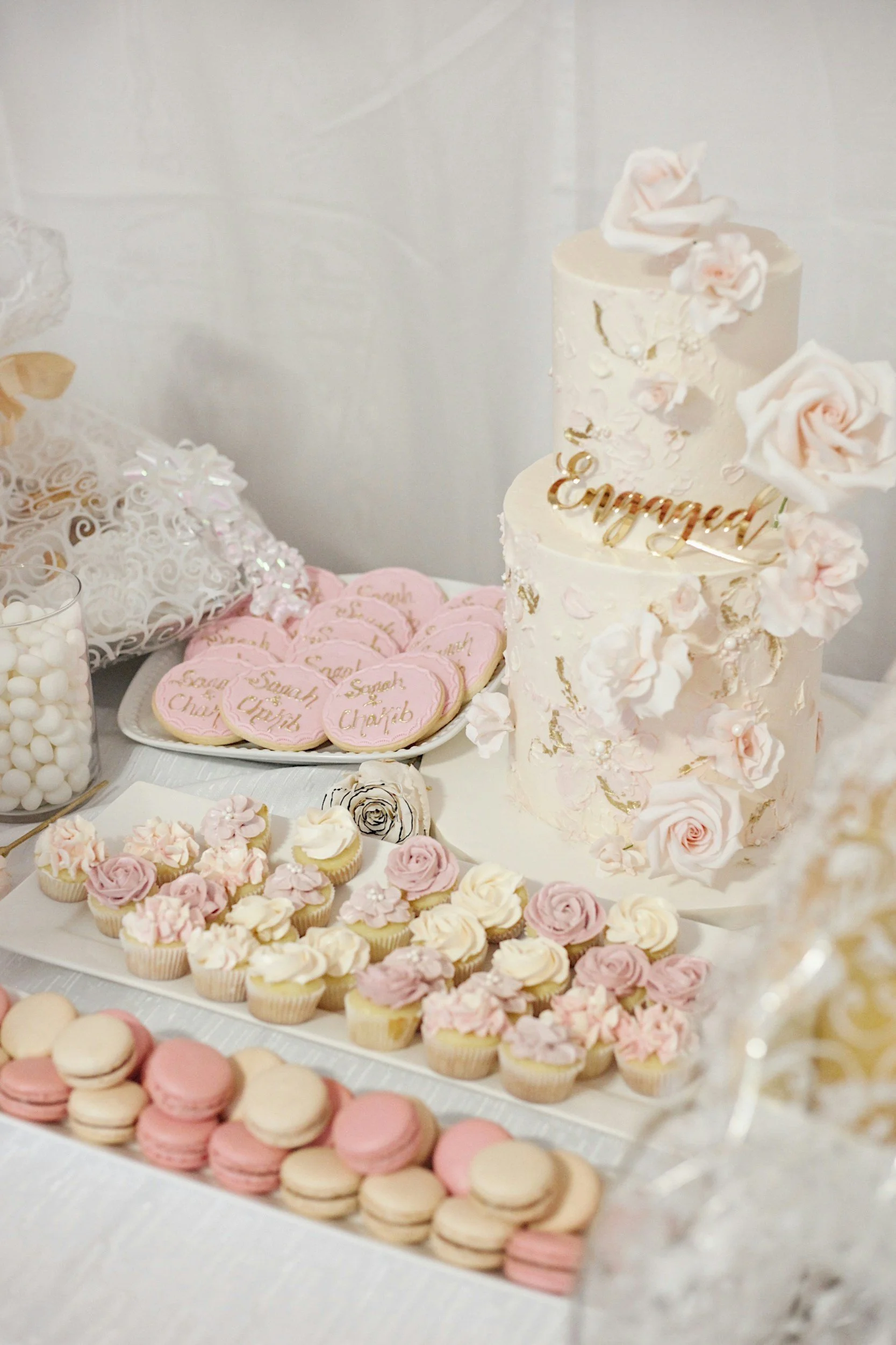 A dessert table featuring a three-tier wedding cake decorated with pink roses and gold accents, with pastel-colored macarons and cupcakes nearby, and pink heart-shaped cookies with gold writing reading 'Sarah & Chayb' on a white plate.