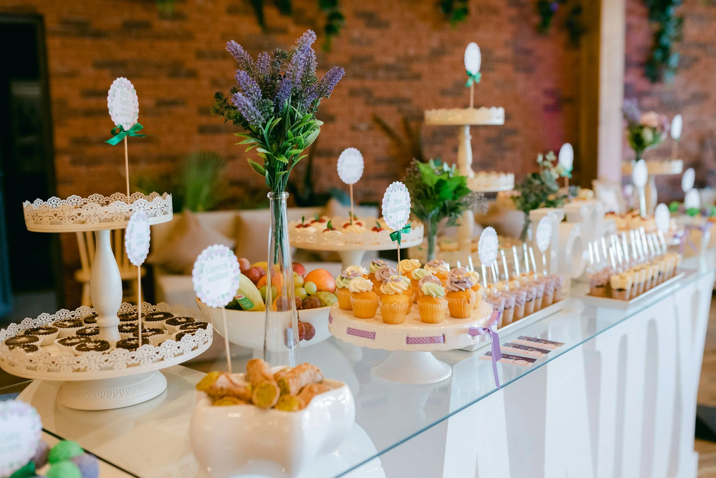 A dessert table featuring cupcakes with swirled frosting, pastries, and fruit, decorated with floral arrangements and small signs, set against a brick wall.