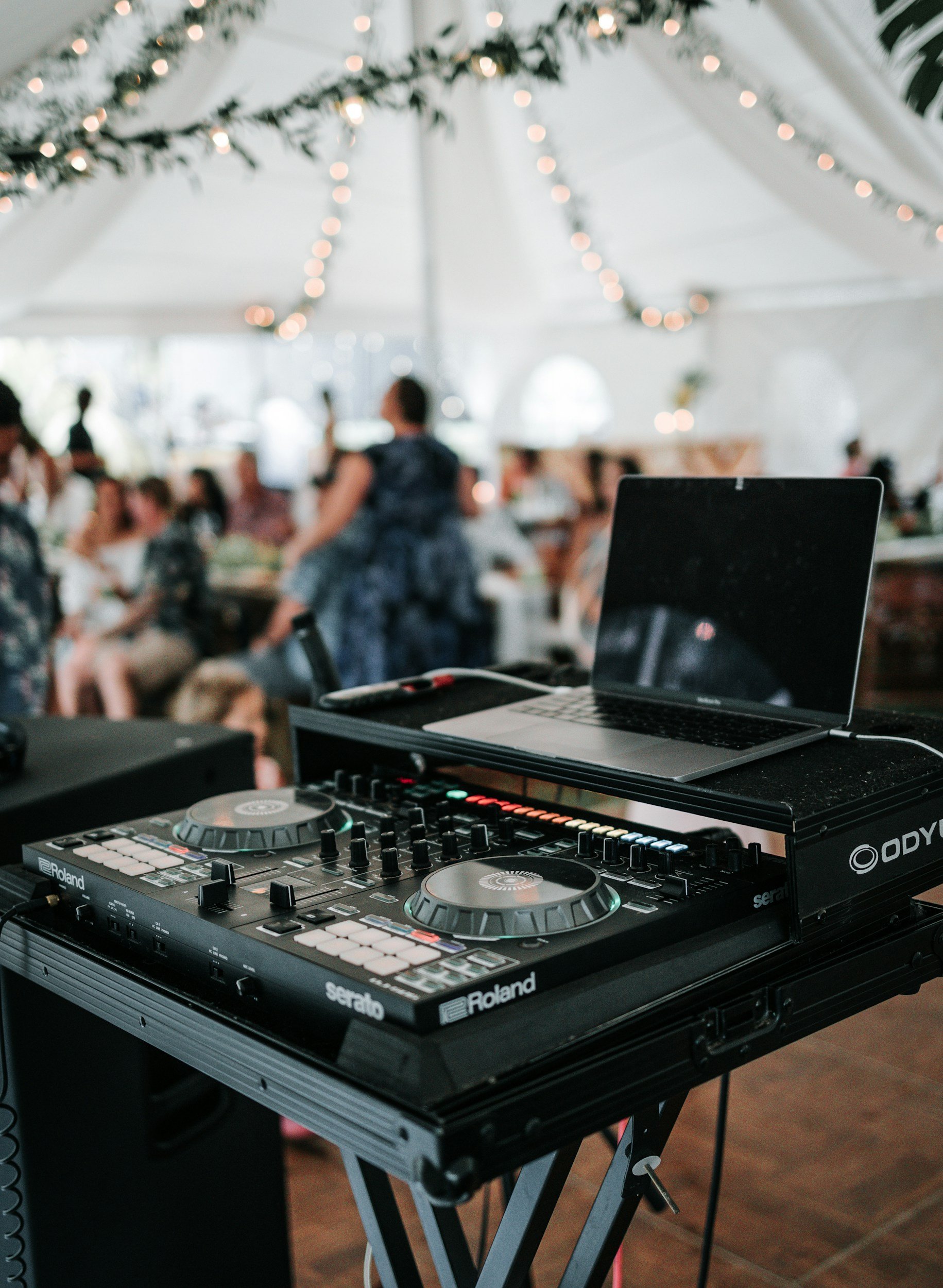 A DJ setup with a Roland Serato controller and a laptop on a black table at an indoor event with white drapes and string lights. There are people in the background, some sitting and some standing.