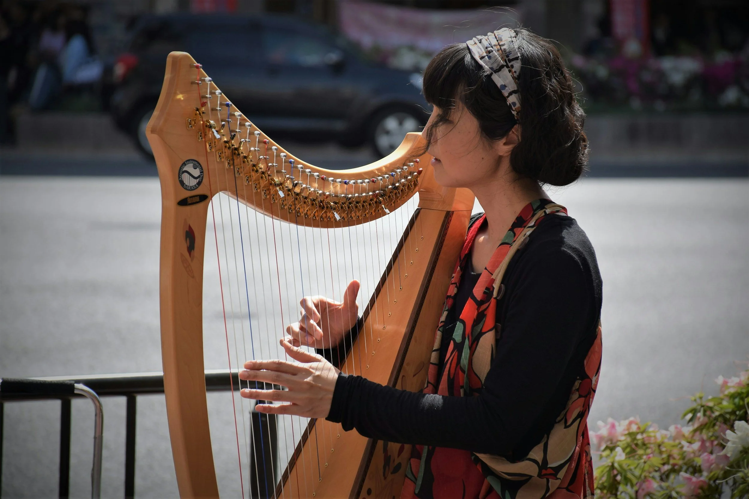 A woman playing a harp outdoors, with cars and people in the background.