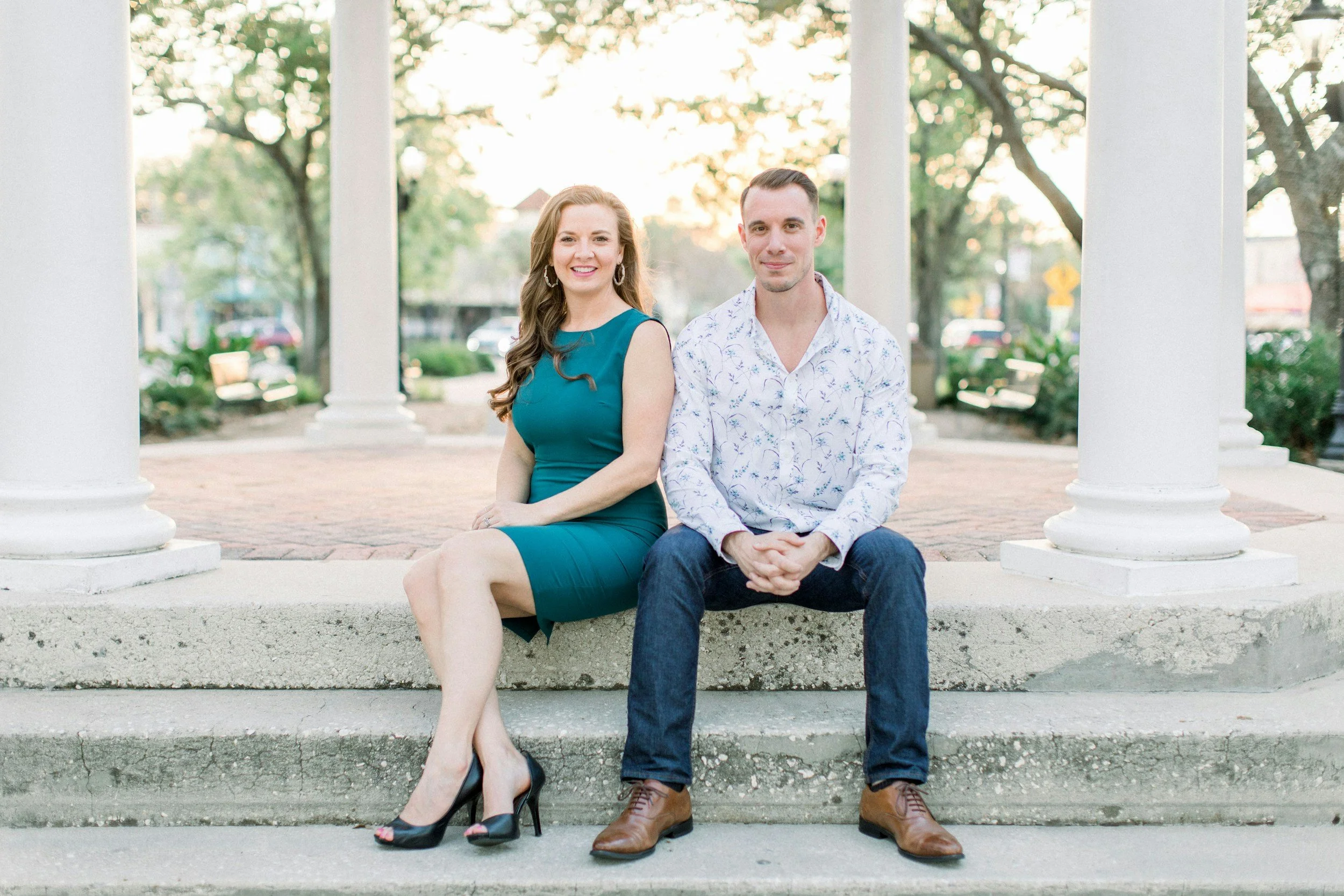 A man and a woman sit on a concrete bench under white columns in a park, smiling at the camera. The woman is wearing a teal dress and black heels, while the man is dressed in a white patterned shirt and dark jeans with brown shoes. Trees and a street with cars are visible in the background during sunset.