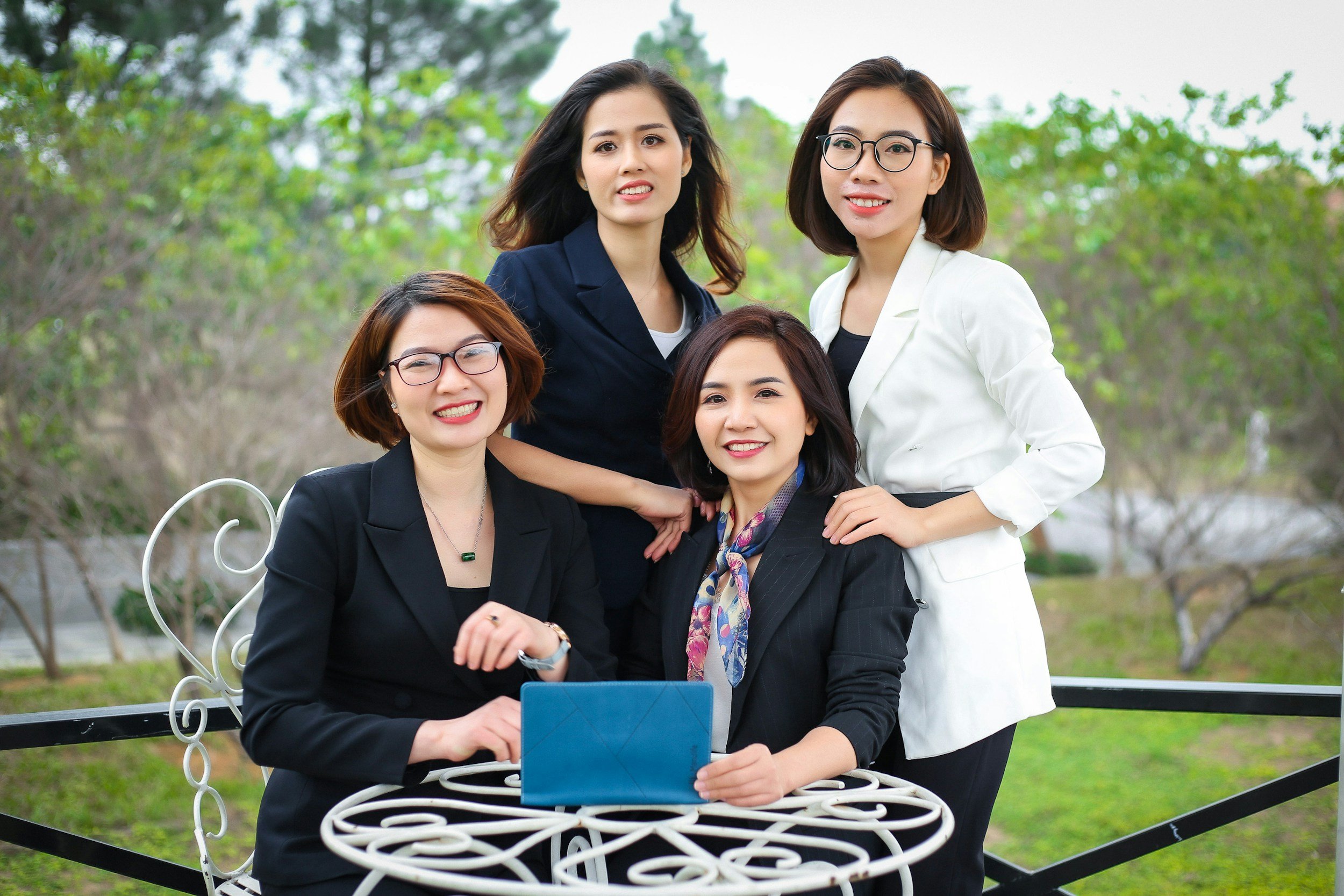 Group of five women in professional attire gathered outdoors in a park, sitting on a white bench with one woman holding a tablet.
