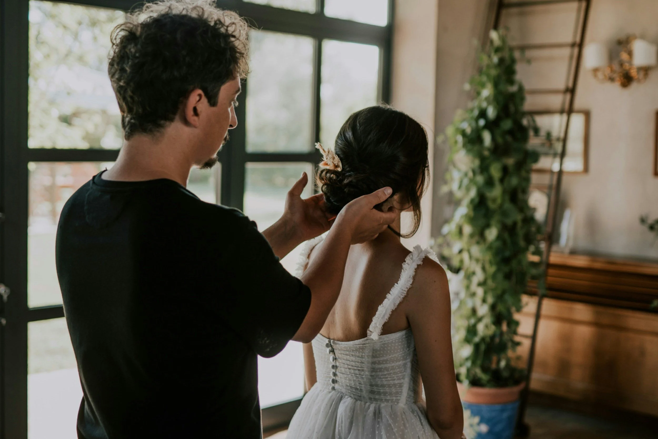A man dressing a woman in a white strapless dress, possibly for a wedding, in an indoor setting with large windows and green plants.