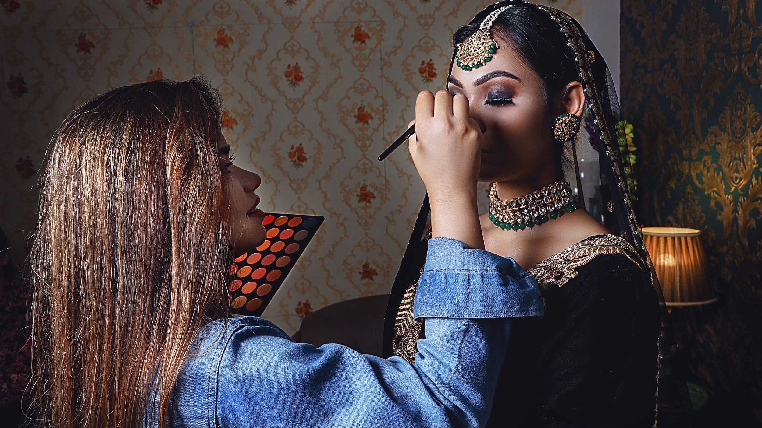 A woman with long hair wearing a denim jacket applying eye makeup to another woman dressed in traditional Indian attire with jewelry.