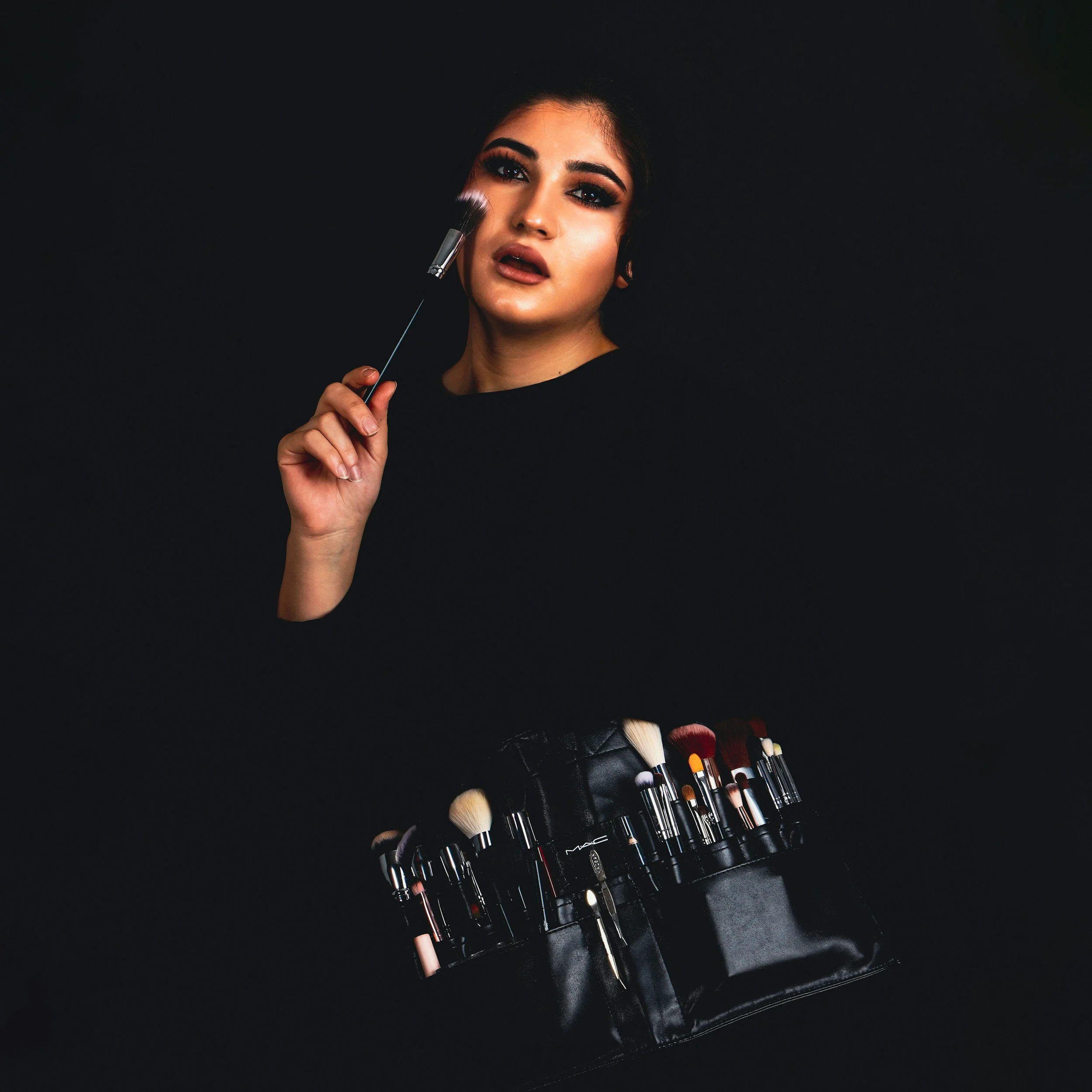 A woman with dark hair and dramatic makeup, holding a makeup brush near her face, with a case of various makeup brushes below her, set against a black background.