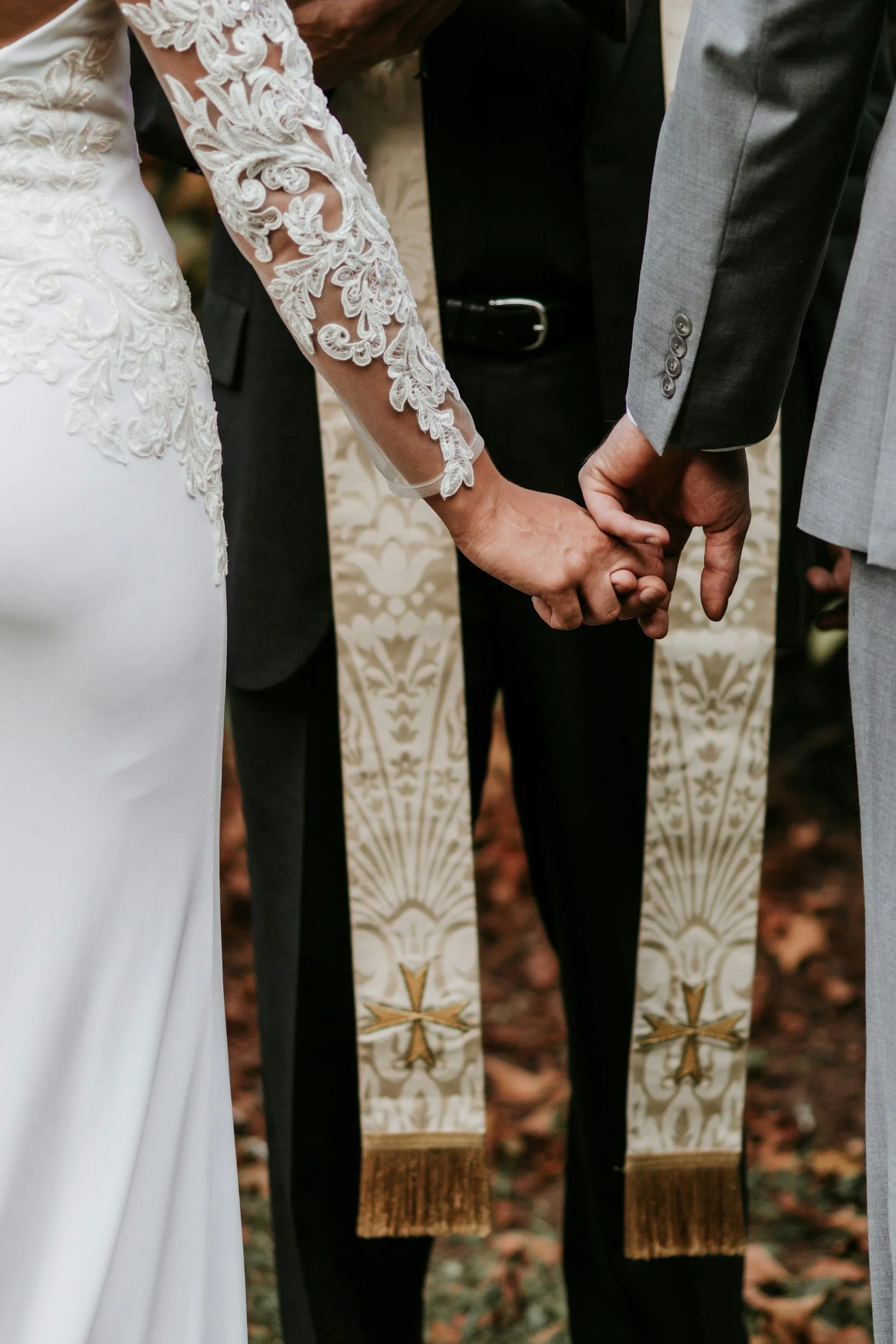 Close-up of a couple holding hands during a wedding ceremony, with the bride wearing a lace wedding gown and the groom in a light gray suit.