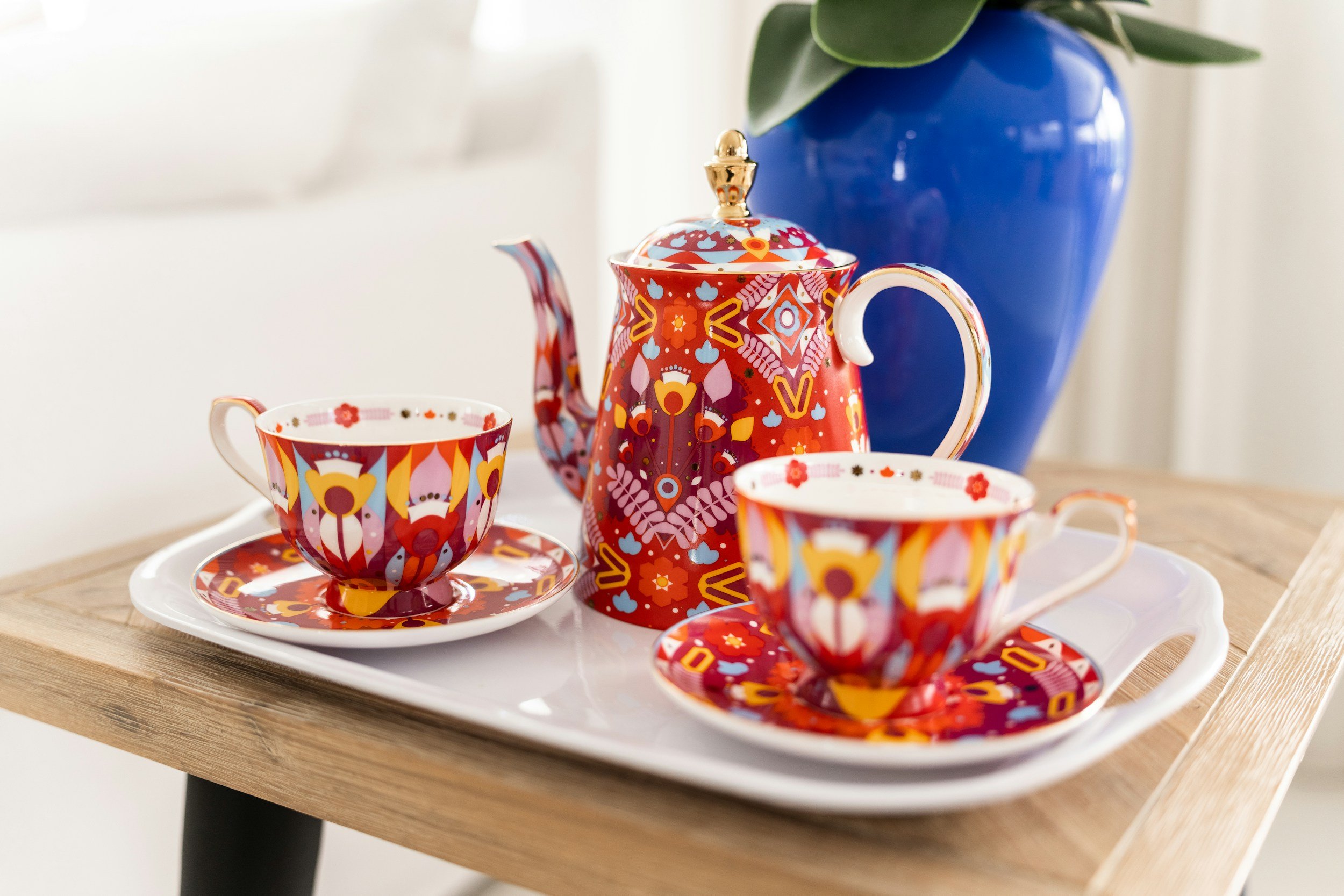Colorful, patterned tea set with matching cups, saucers, and a teapot on a tray, with a blue vase and greenery in the background.