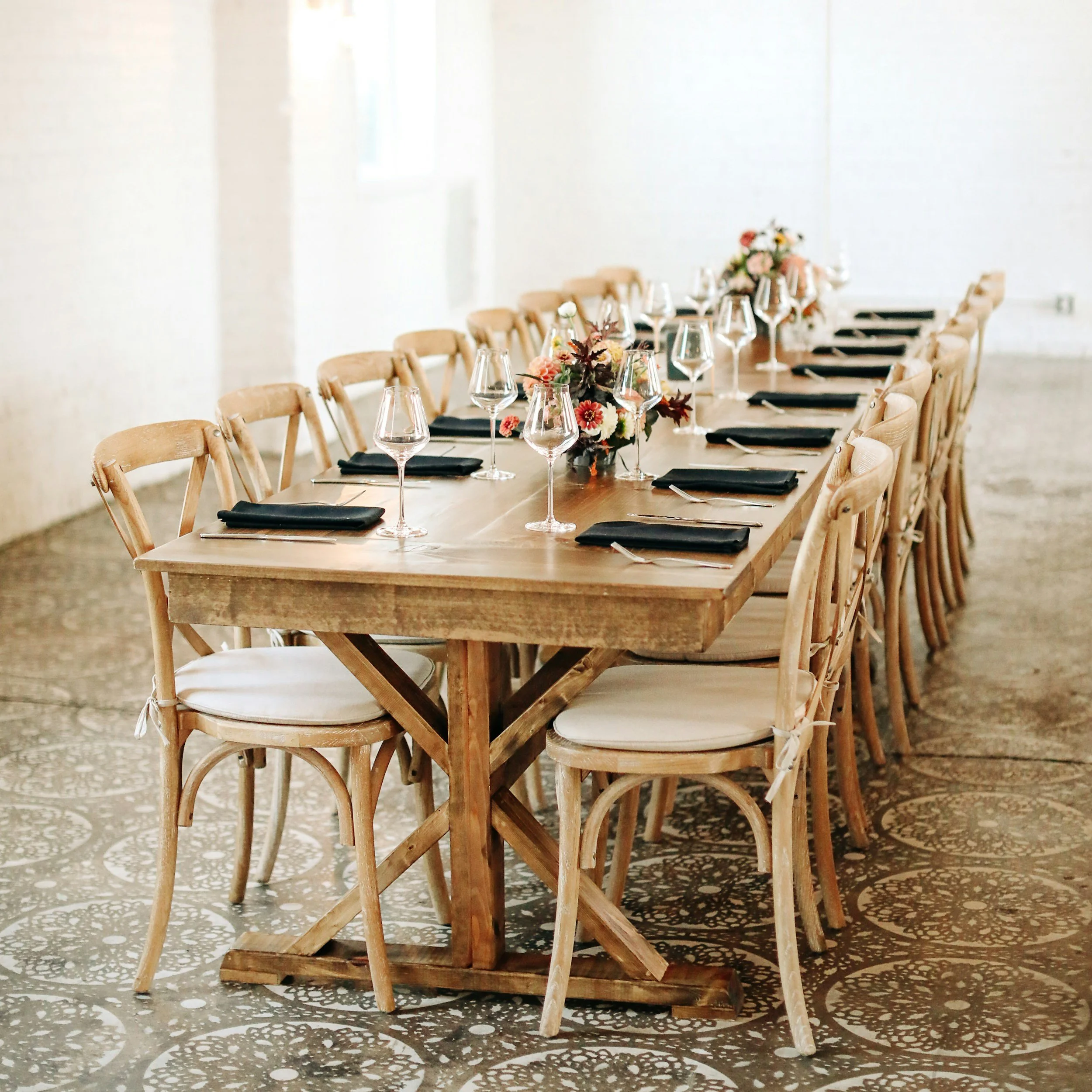 A long wooden dining table set with black napkins, wine glasses, and floral centerpieces in a bright room with patterned tile floor and white walls.