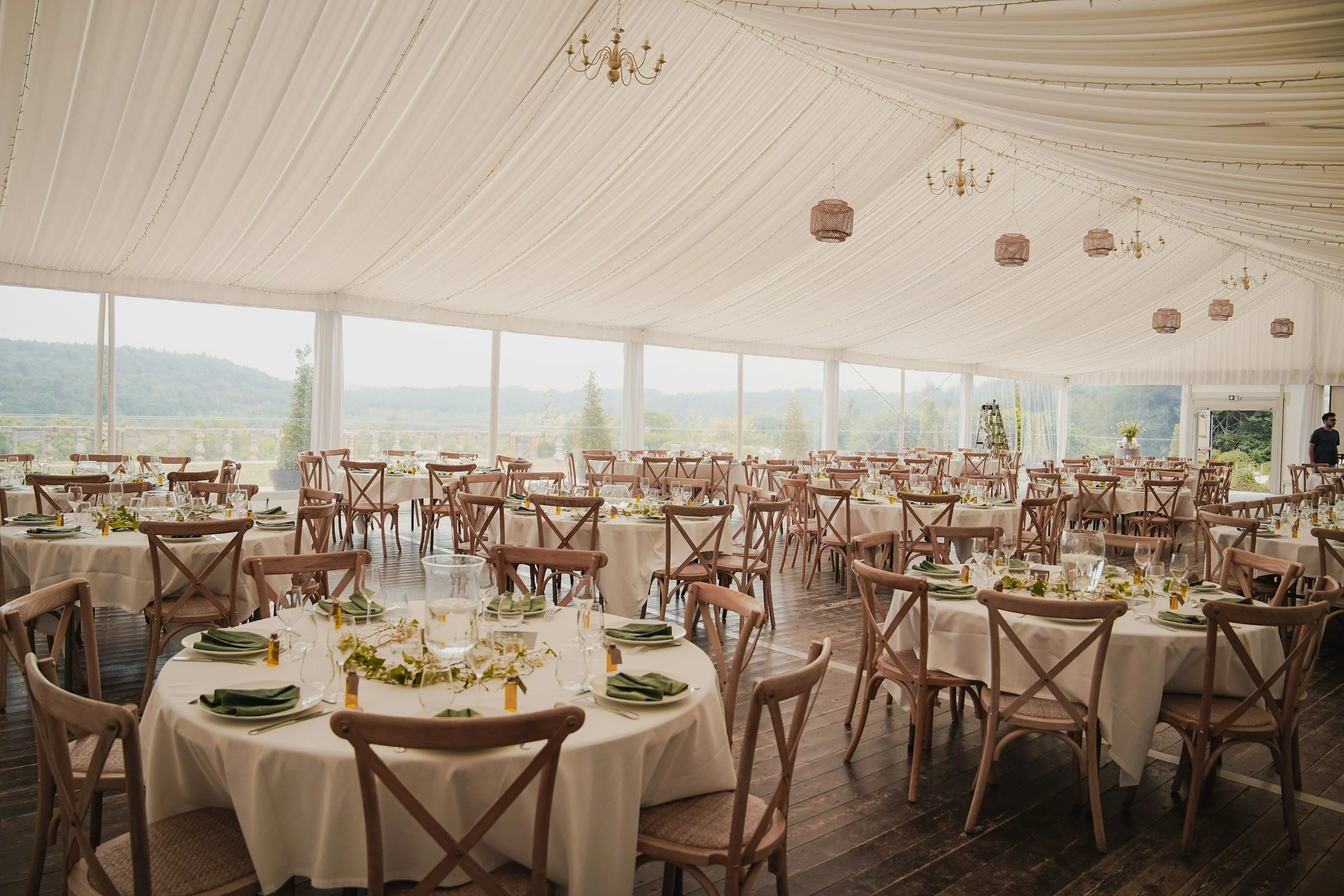 Large white tent interior set up for an event with round tables, green napkins, glassware, and wooden chairs. Chandeliers and pink hanging lamps decorate the ceiling. Large windows offer a view of distant hills and trees.