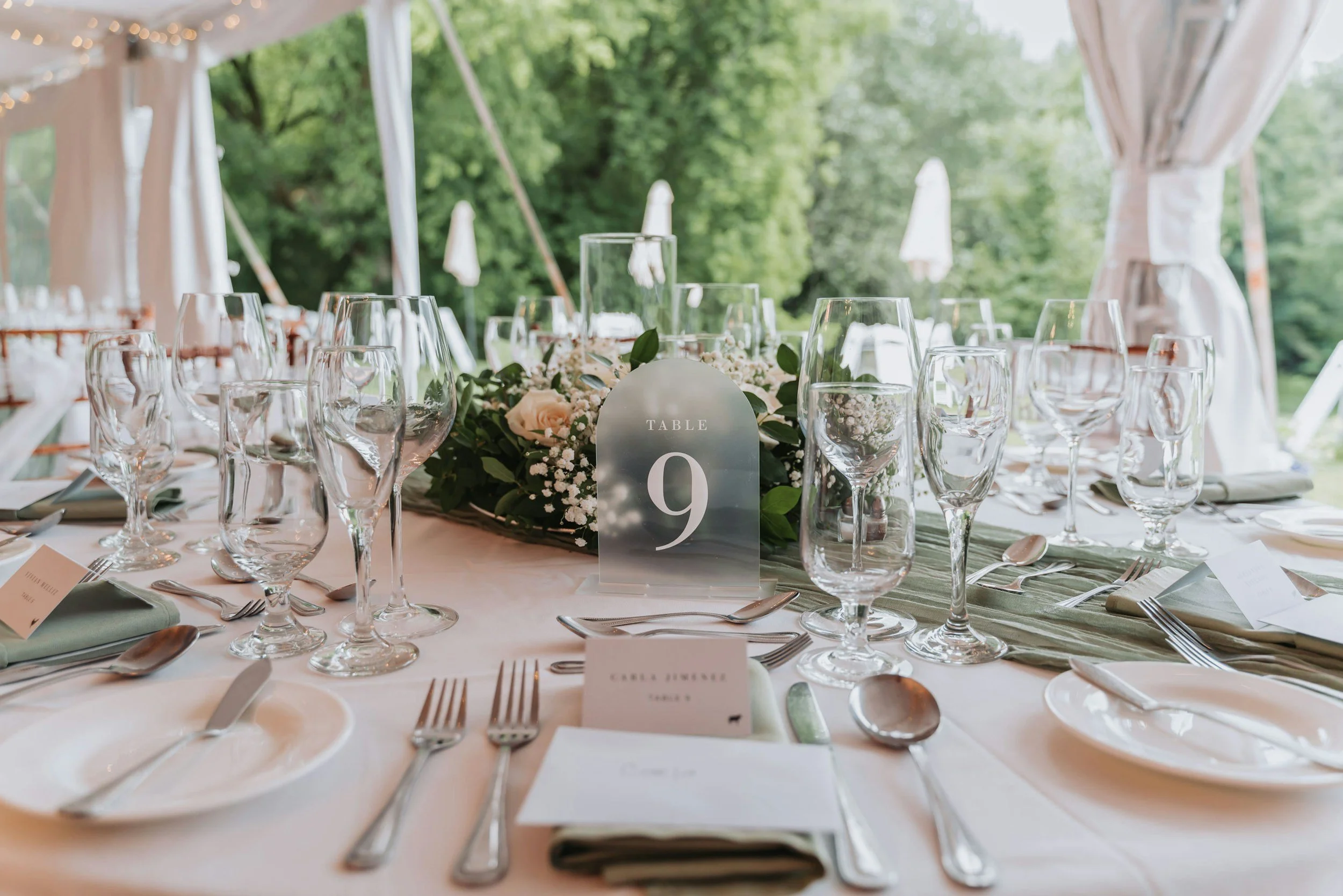Elegant outdoor wedding reception table set with glassware, silverware, white napkins, floral centerpiece, and a table number sign in a tent with greenery outside.