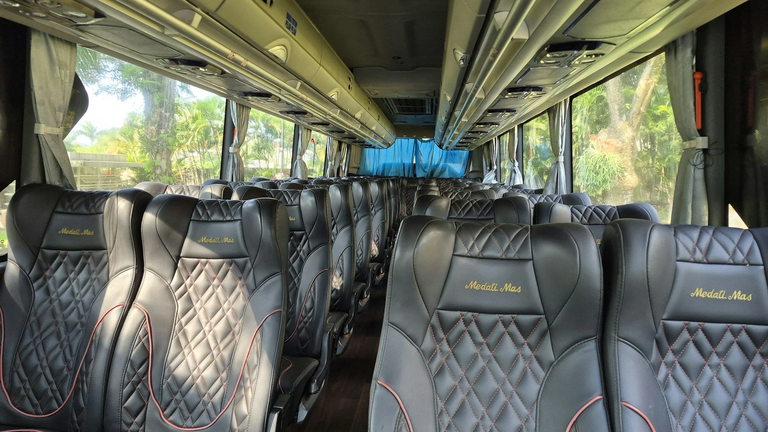 Interior of a bus with black leather seats, large windows with curtains, overhead compartments, and a view of lush green trees outside.