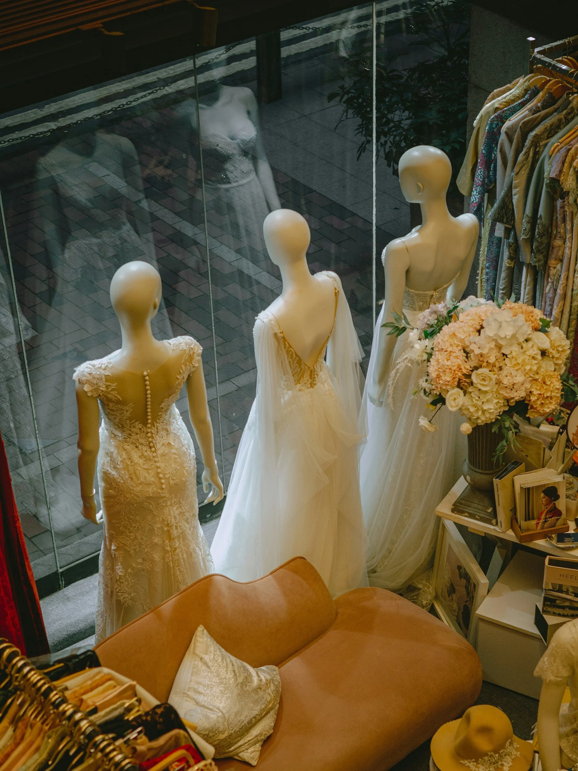 Display window with three mannequins dressed in wedding gowns and a collection of dresses on hangers. Mannequins are seen through a glass window, with reflections of a woman outside and a plant in the background. There's also a bouquet of flowers and a shelf with books inside the store.