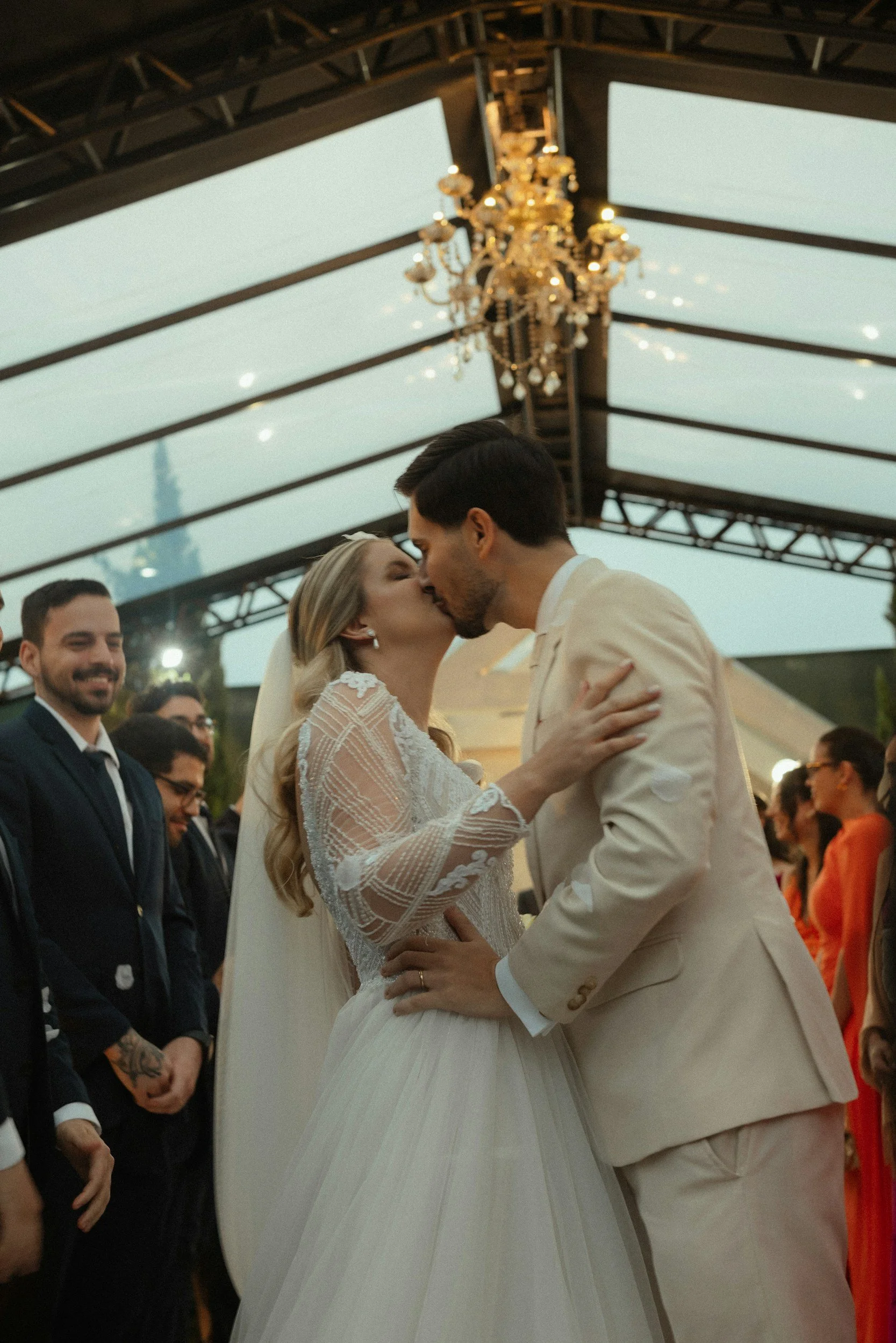 A bride and groom kiss during their wedding ceremony under a decorated outdoor pavilion, with guests and a chandelier overhead.