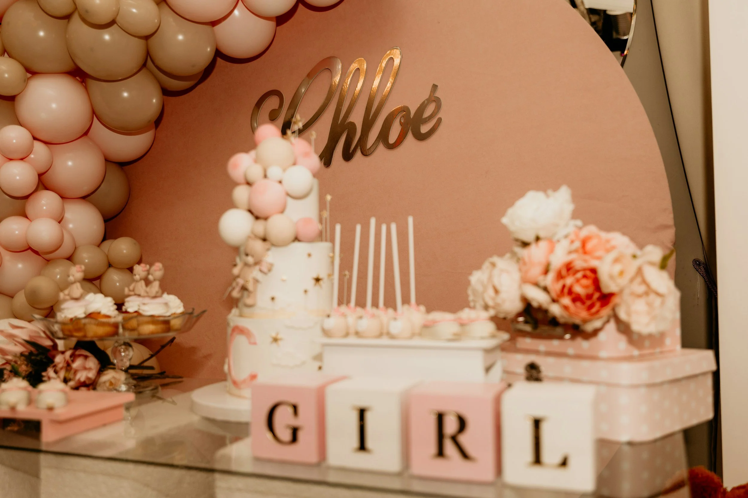 Decorative party table with pink themed balloons, flowers, cookies, and a cake. There is a sign that reads 'Chloe' in gold letters above the table.