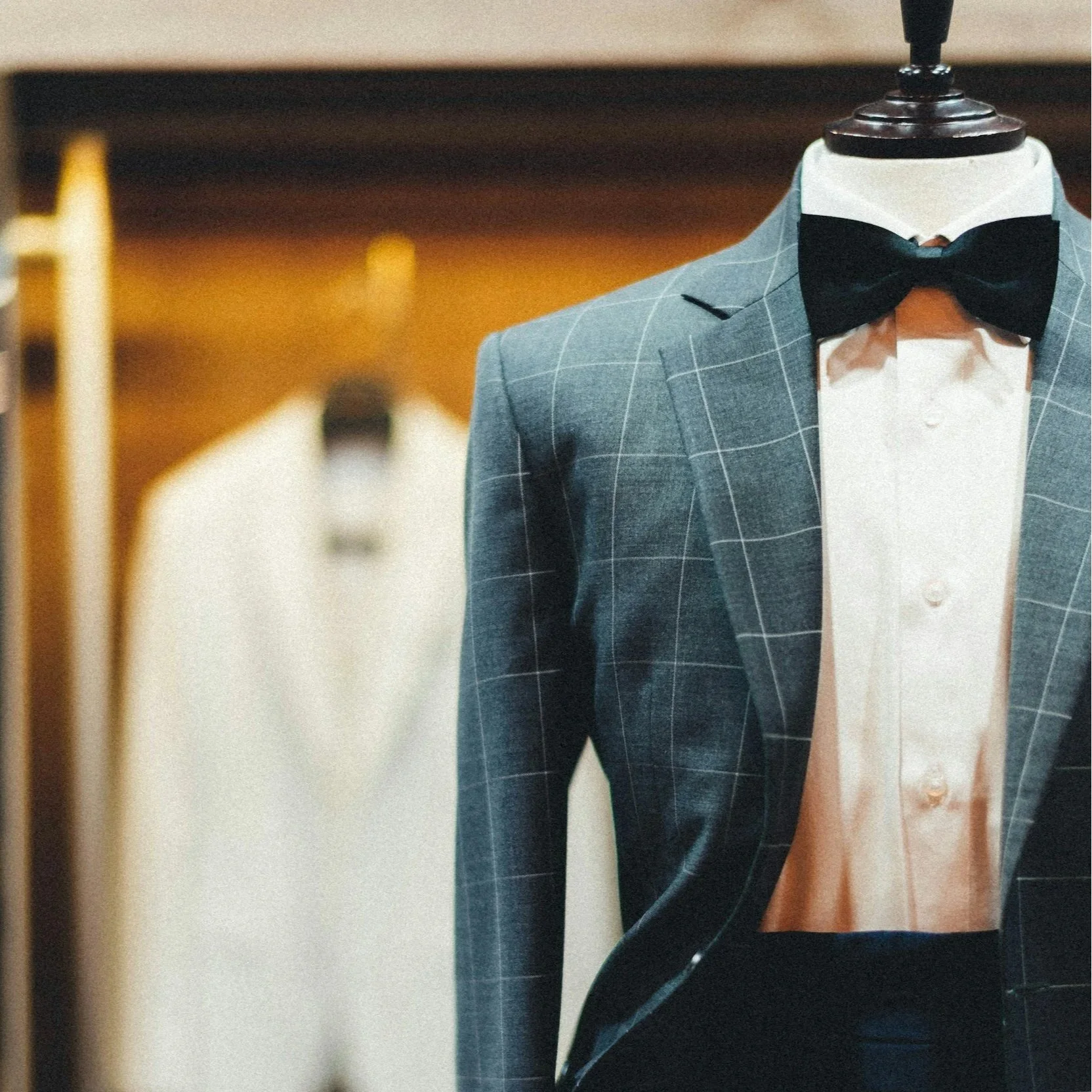 A tuxedo jacket with a checkered pattern, a white dress shirt, and a black bow tie on a mannequin, inside a clothing store or dressing room.