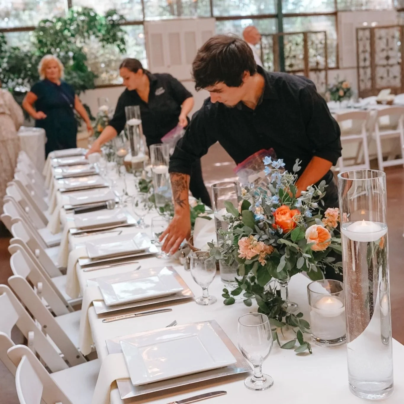 People preparing a long banquet table with white tablecloth, floral centerpieces, and candle decorations in a well-lit indoor space.