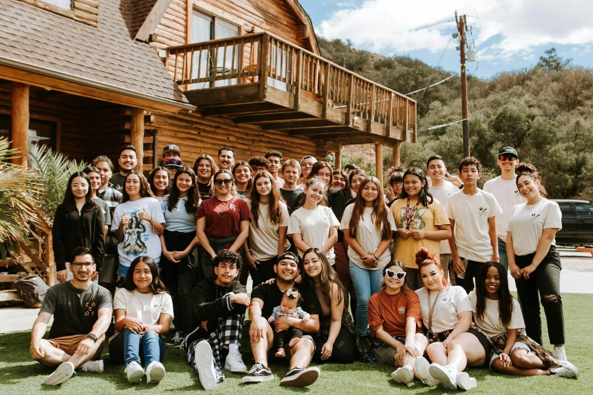 A large group of young people and a few adults posing in front of a rustic wooden house with a mountain background. Everyone is smiling.