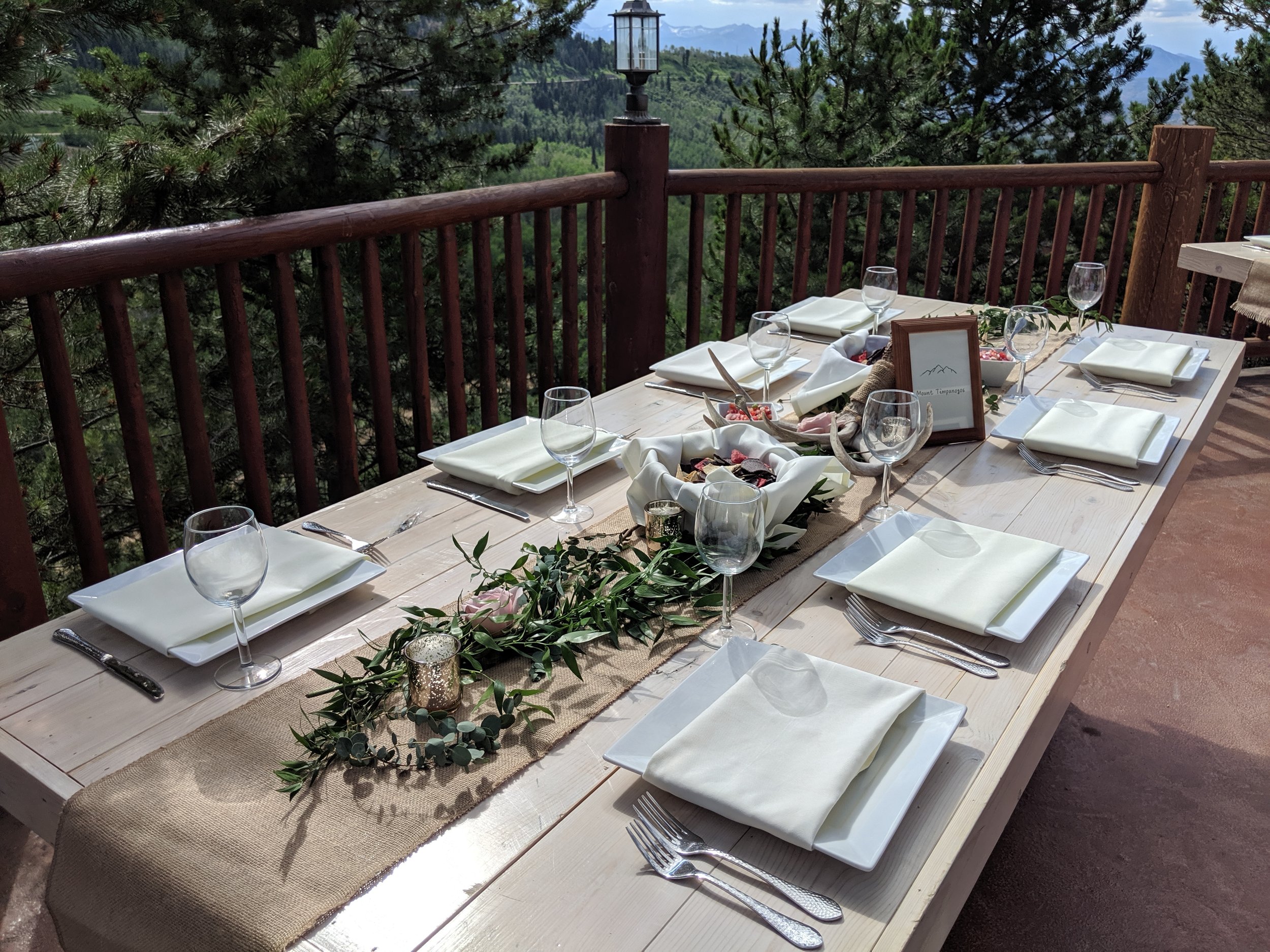 An outdoor dining table set for dinner on a wooden deck with a mountain view in the background. The table is decorated with greenery, candles, and framed signs, and is prepared with white plates, silverware, and wine glasses.