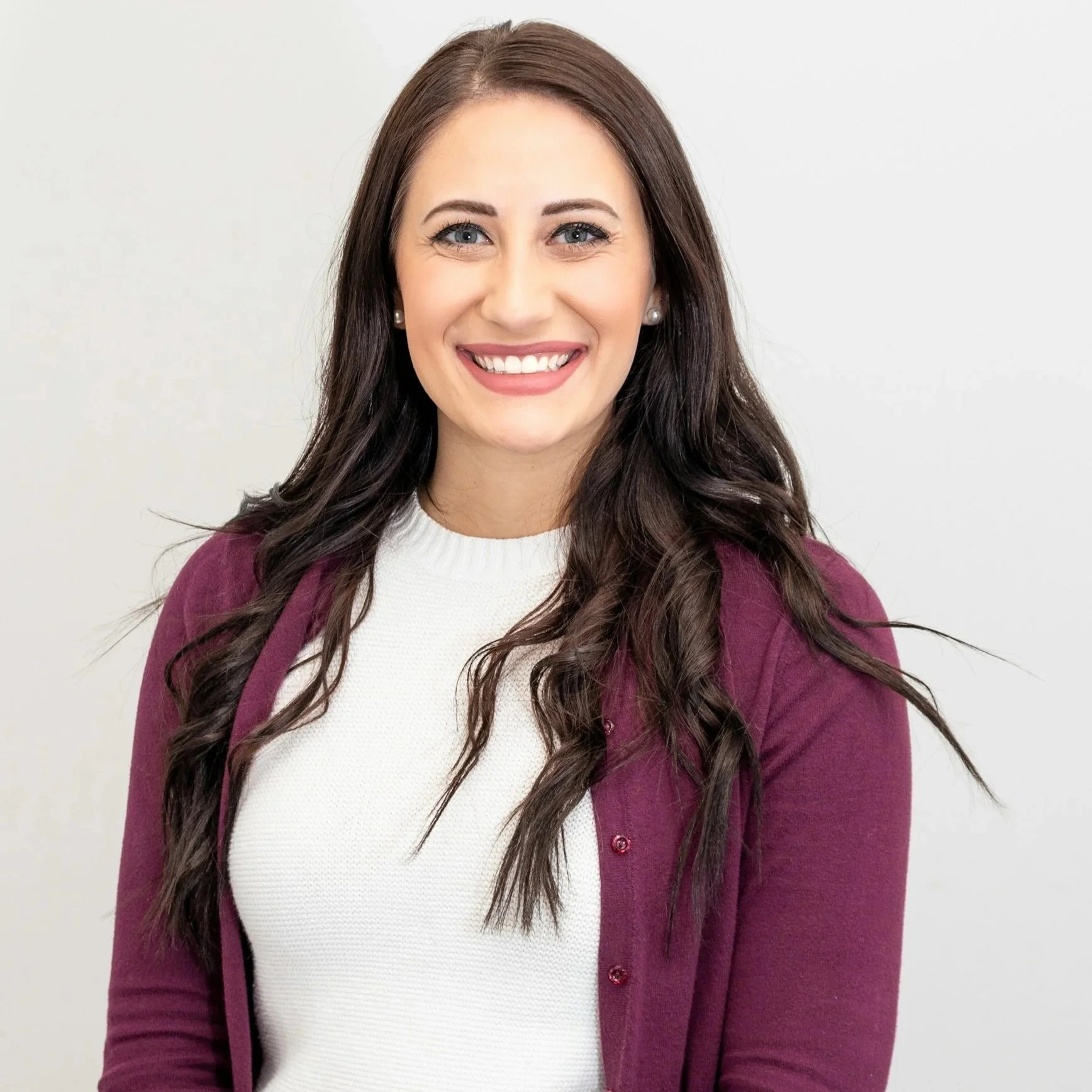 Portrait of a woman with long brown hair, blue eyes, smiling, wearing pearl earrings, a white top, and a maroon cardigan, against a plain white background.