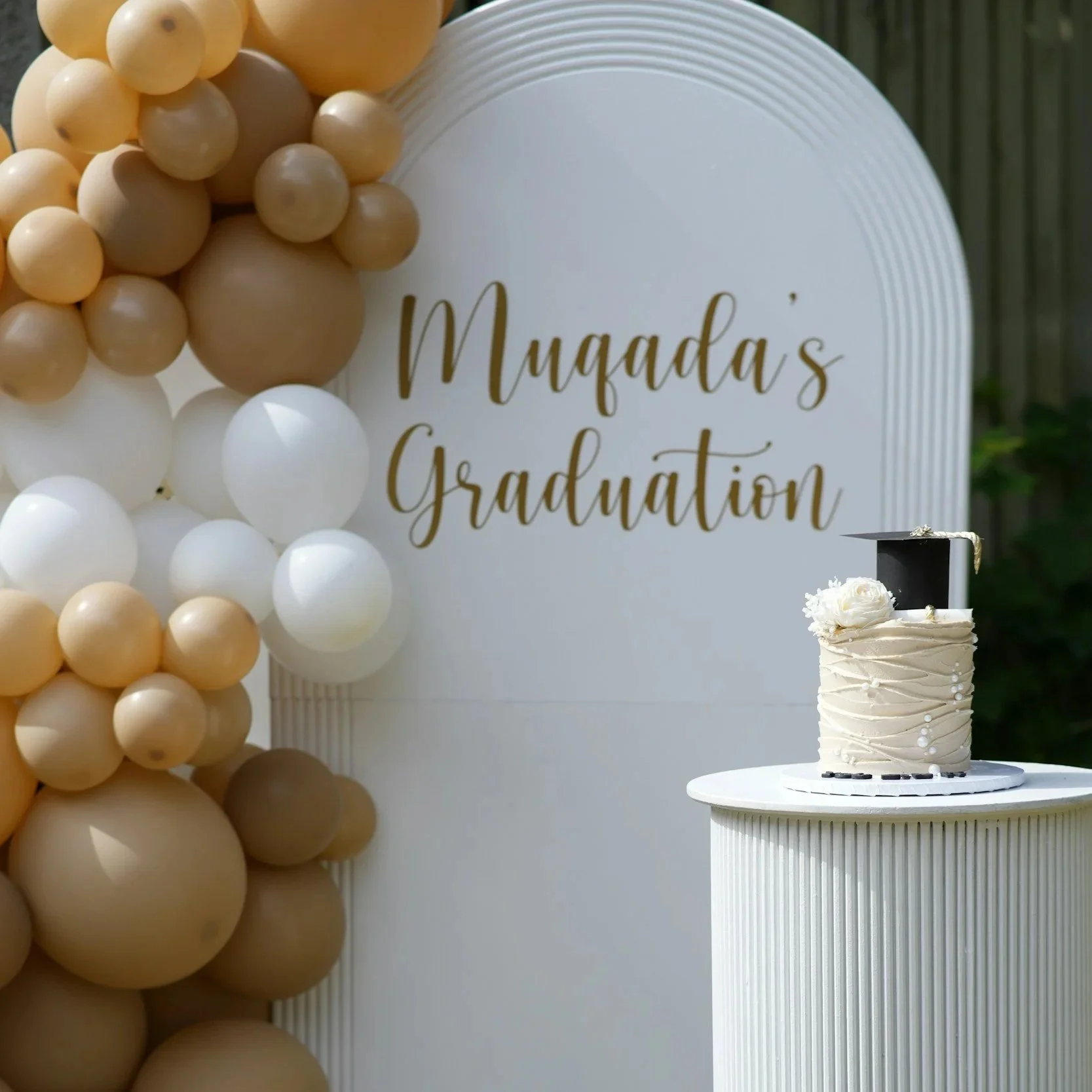 Celebration setup with beige and white balloon arch, white circular backdrop with gold lettering that reads 'Muada's Graduation', and a white tiered cake with floral decoration on a pedestal