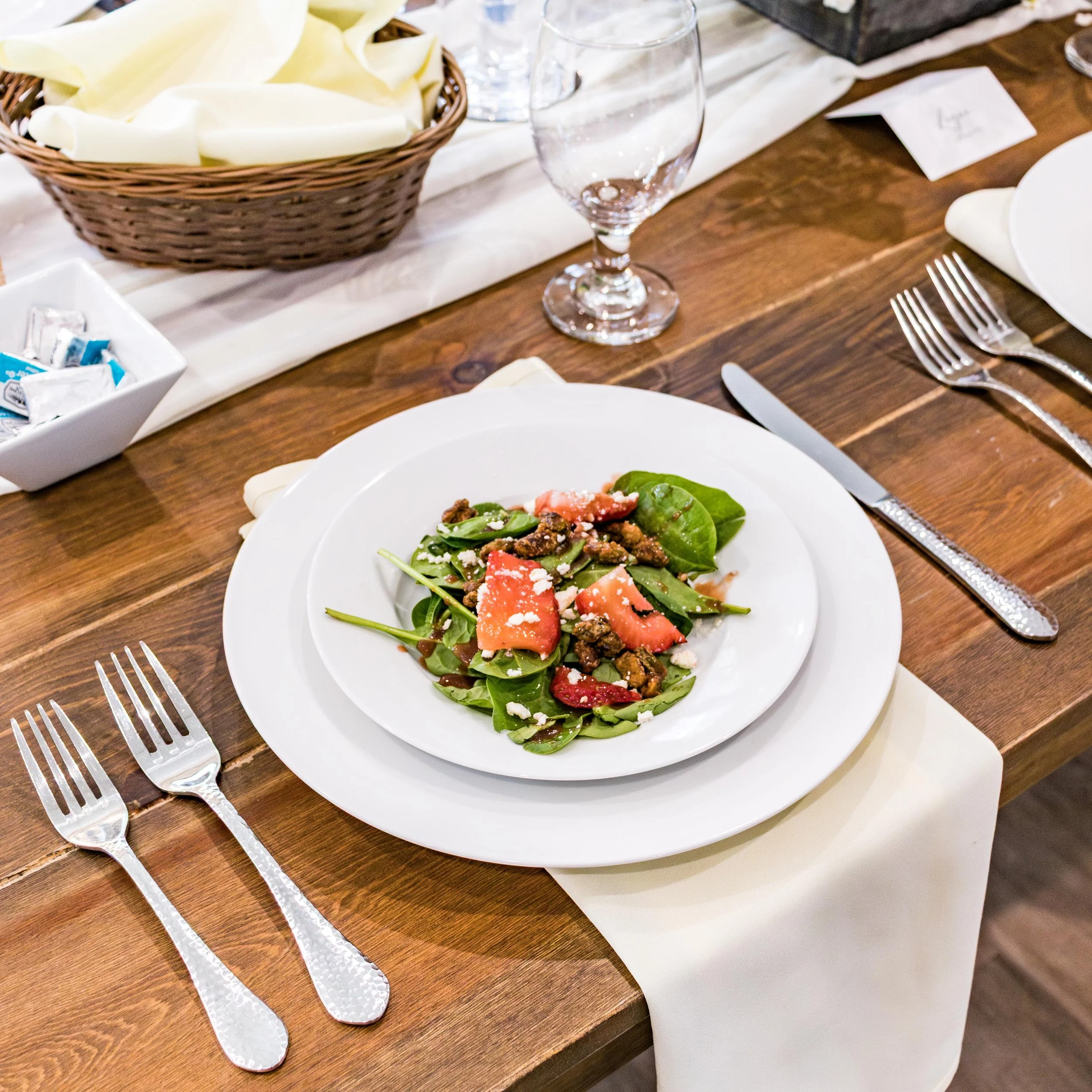 A wooden dining table set for a meal with white plates, silver forks, and cloth napkins. There is a dish of salad with greens, tomatoes, and cheese, a basket of sliced cheese, and a glass of water.