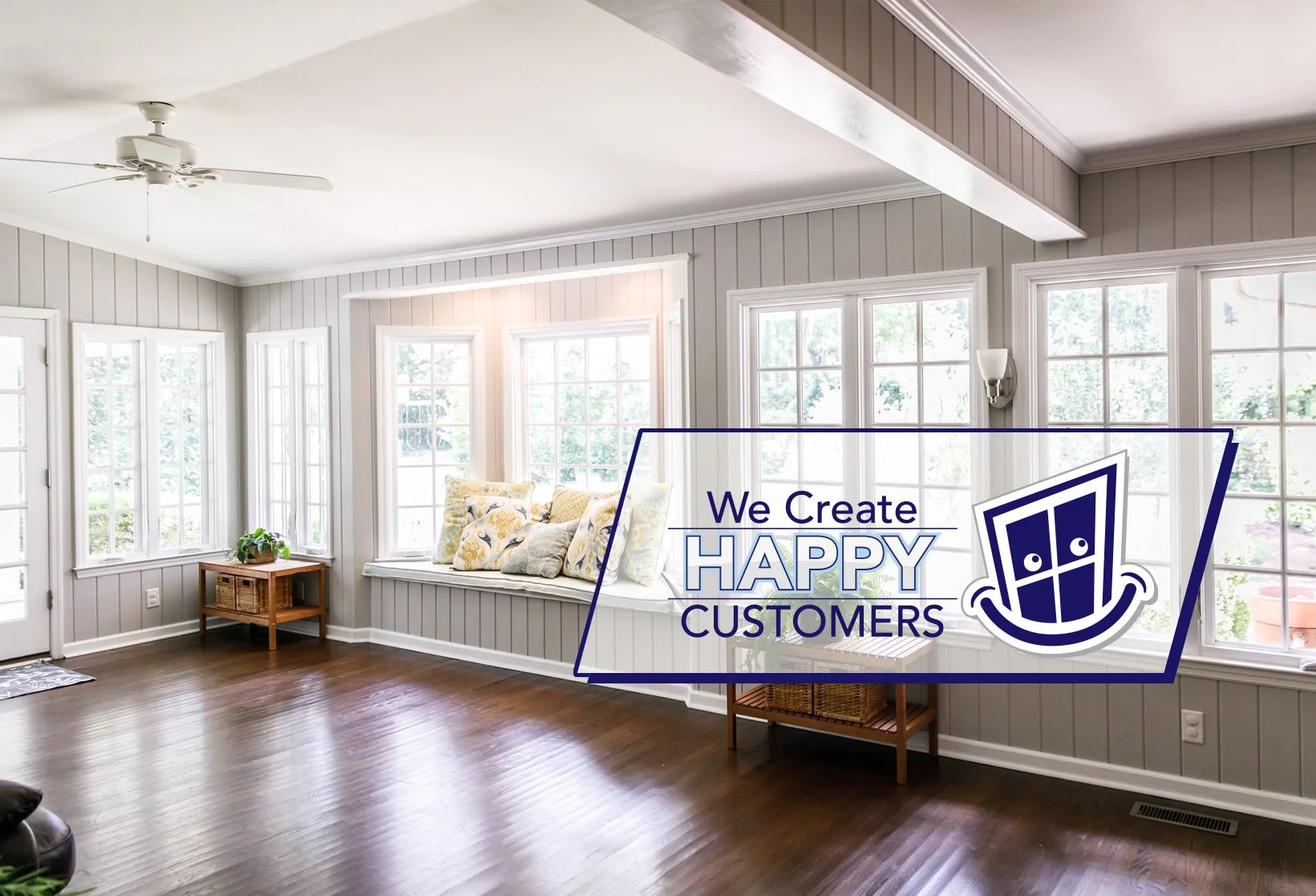 Bright living room with large windows, white walls, hardwood floors, a white ceiling fan, and a window seat with pillows. A small wooden table with a potted plant is near the corner. An overlay graphic reads 'We Create Happy Customers' with a logo.
