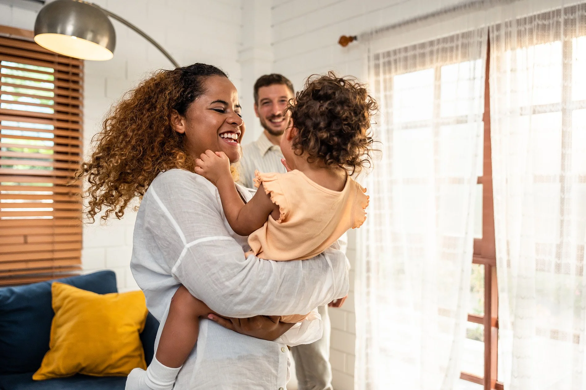 A woman is holding a young girl in her arms while a man looks on and smiles at them in a brightly lit room with curtains and a couch with yellow and blue pillows.