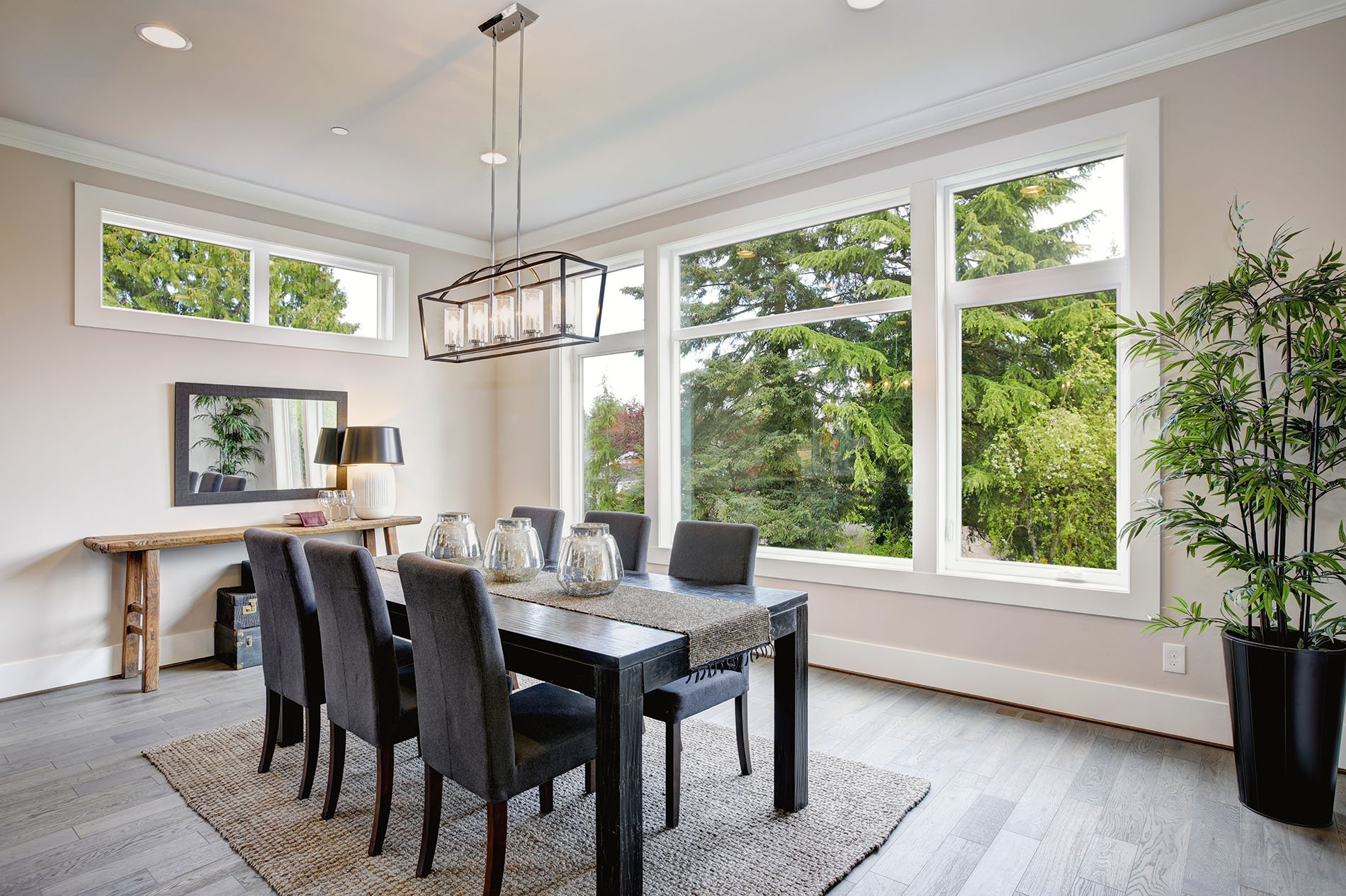 A dining room with a large window showing green trees outside. It has a black dining table with eight gray chairs, a light wood console with a mirror and lamp, glassware on the table, and a potted plant in the corner. Modern chandelier hangs above the table.
