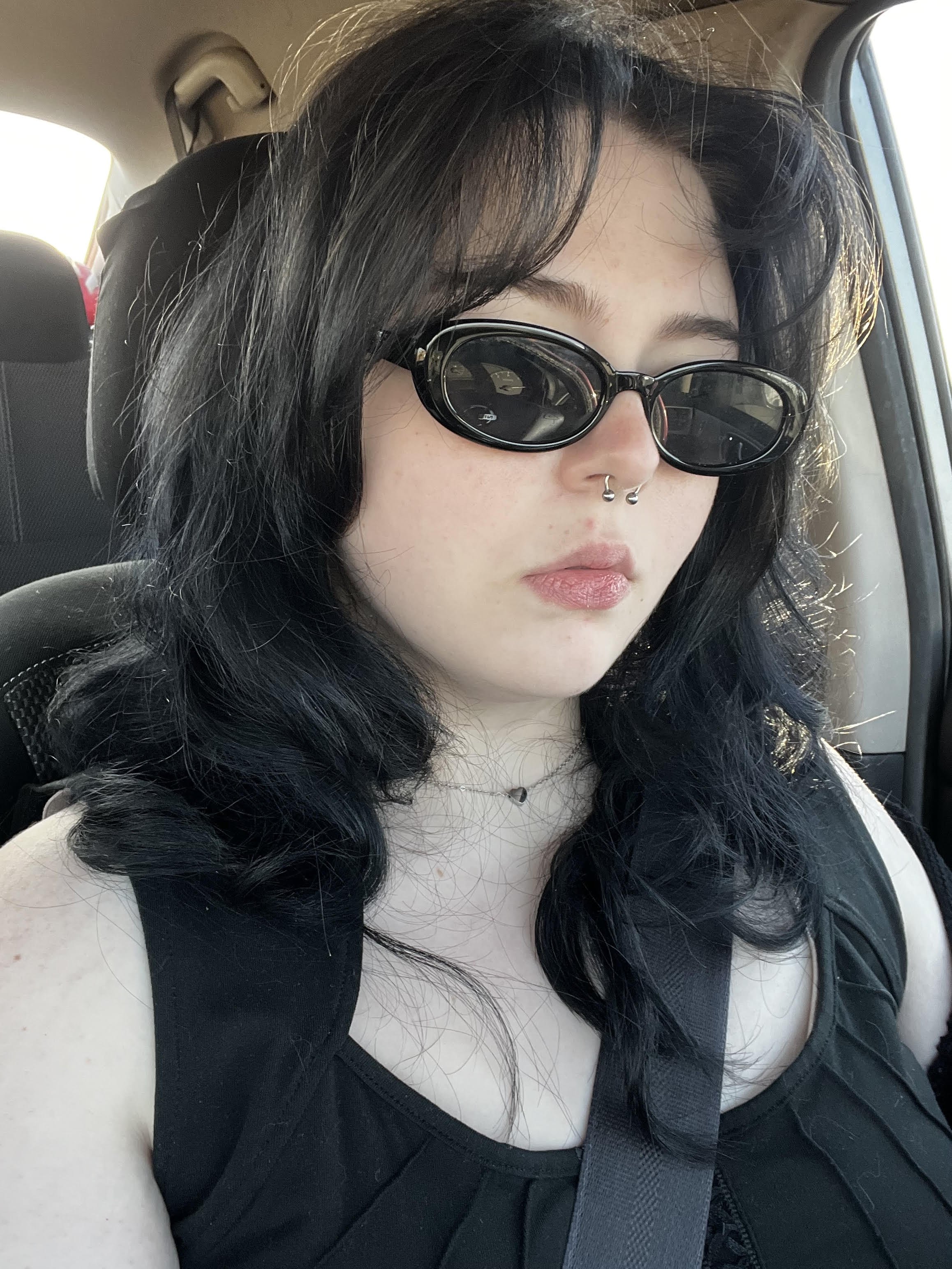 A young woman with dark, curly hair and light skin sitting in a car. She is wearing black oval sunglasses, a nose ring, and a black top, with a silver heart necklace visible. She appears to be looking down or away, with sunlight illuminating part of her face.