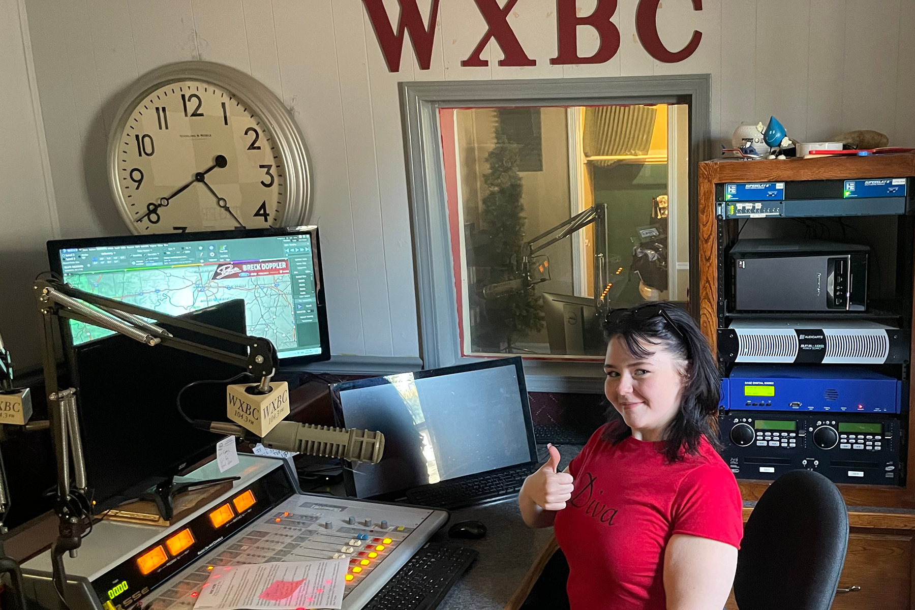 A radio studio with a large wall clock and red letters W X B C on the wall, a woman in a red shirt sitting at a desk with broadcasting equipment, smiling and giving a thumbs-up.