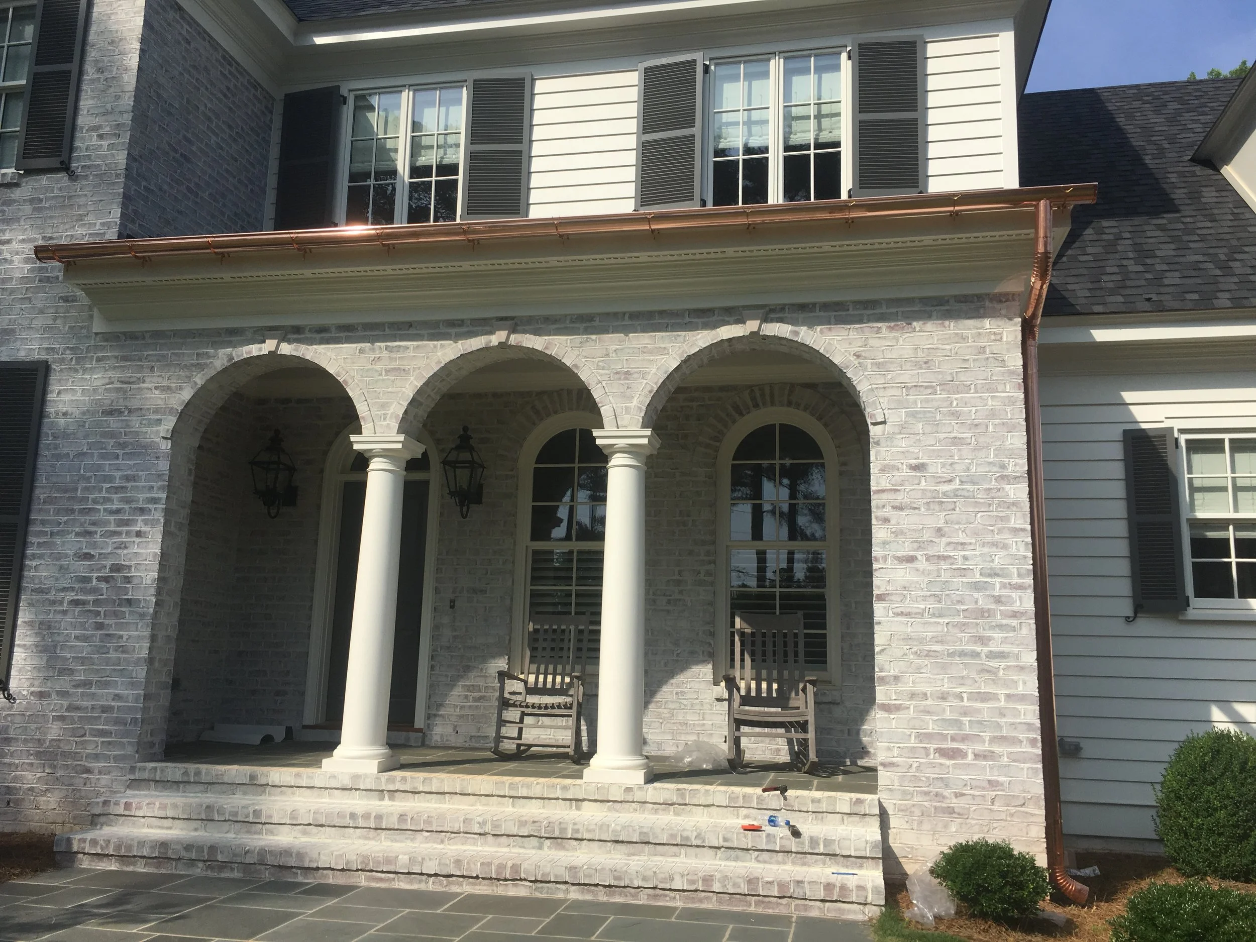 Front view of a two-story house with a brick and siding facade, arched entryway with white columns, and a small porch with two chairs. The house has multiple windows with black shutters and a copper gutter system.