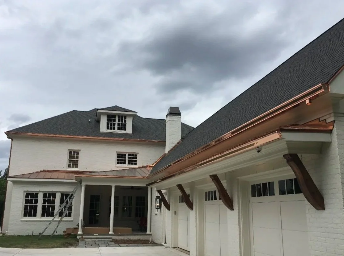 The image shows a large white house with a dark gray roof and copper accents. The house has multiple windows, a small garage door, and a covered front porch with white columns. There are wooden brackets supporting the roof overhang. The sky above is cloudy, indicating an overcast day.