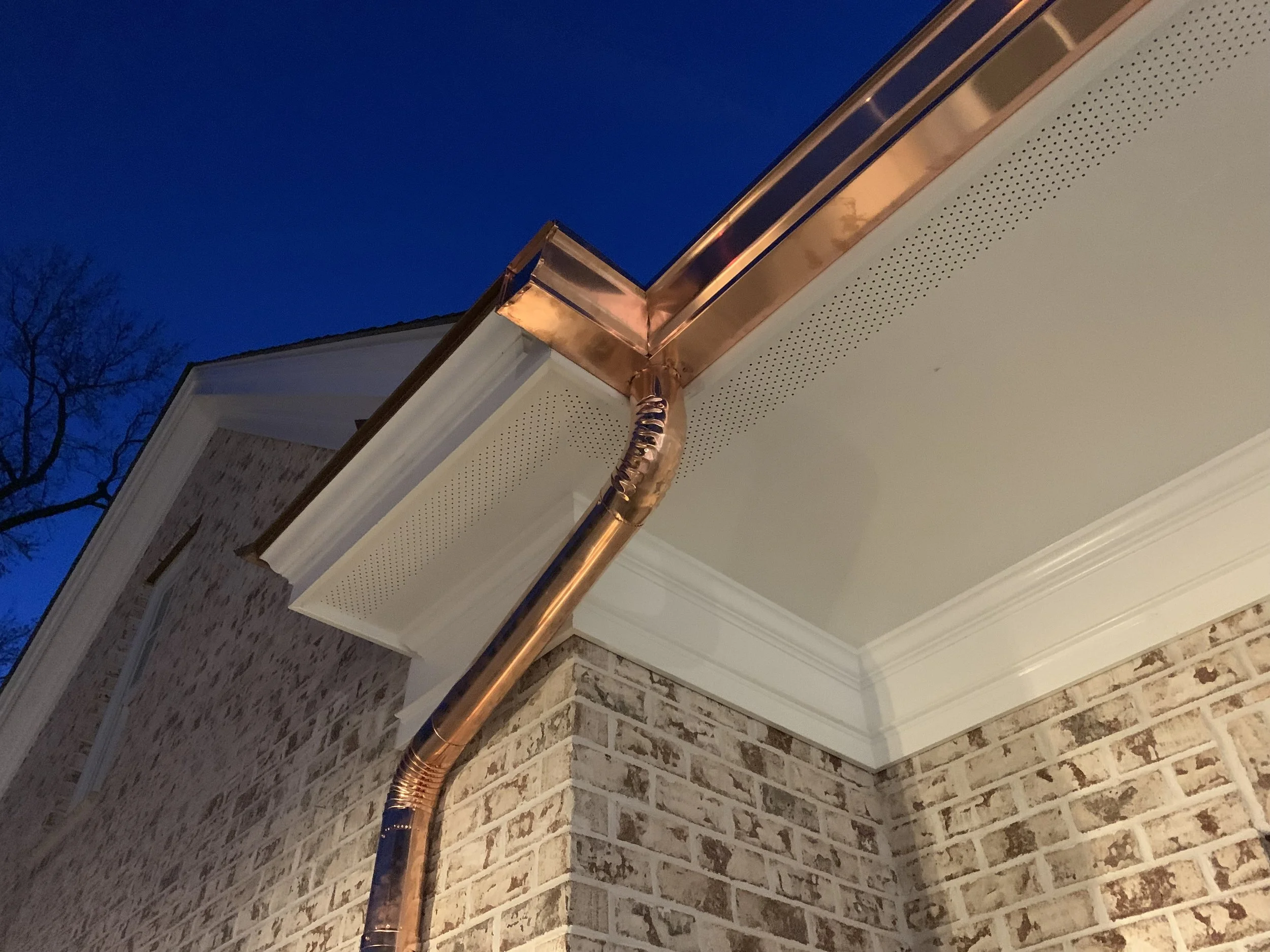 The image shows the corner of a house with beige brick siding, white trim, and a beige soffit with small perforations. A copper-colored metal downspout runs along the corner, connecting to the gutter. The background features a dark blue sky and a leafless tree.