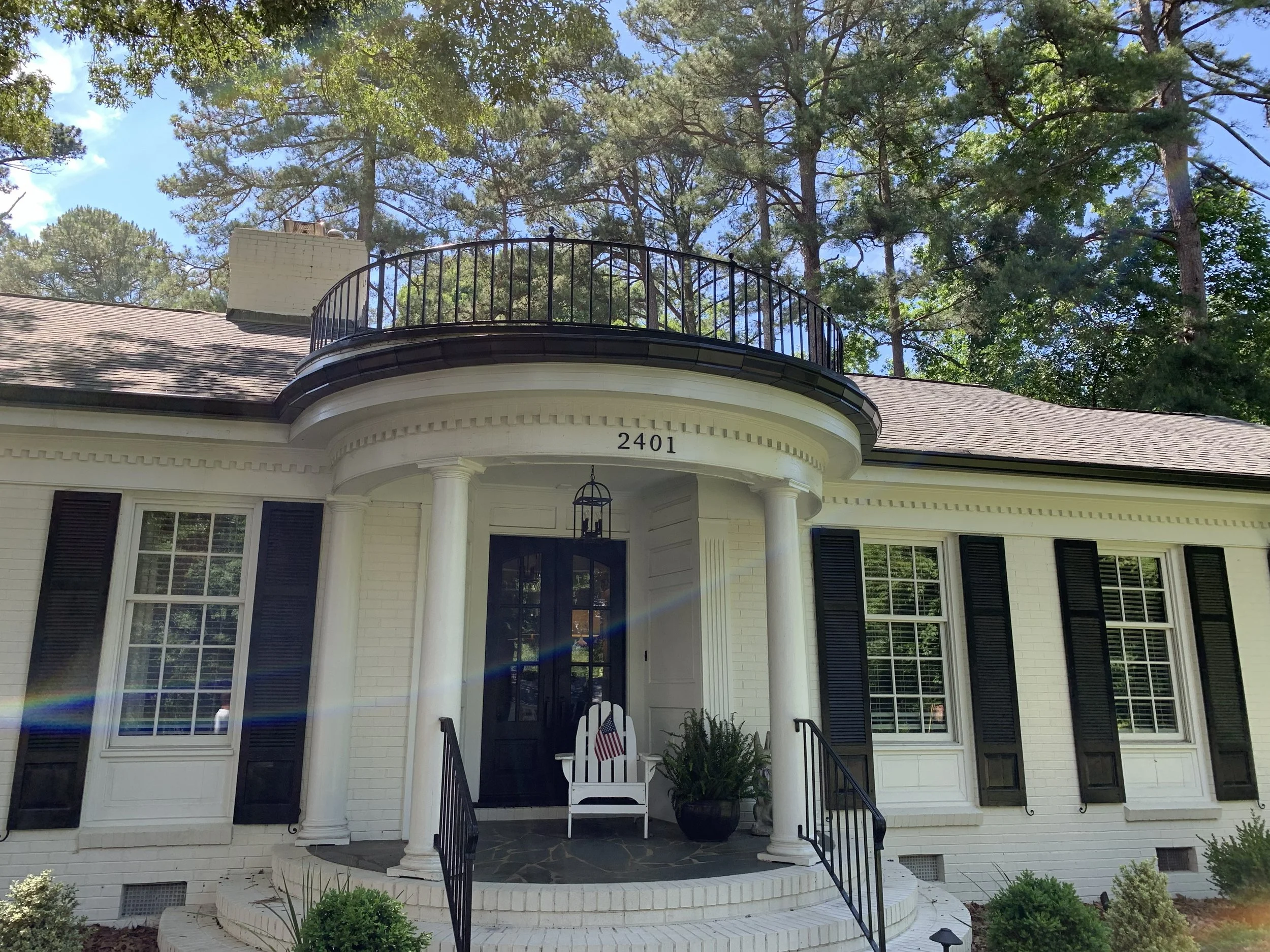 Front view of a white house with black shutters, a rounded porch with white columns, and a black metal railing. A white chair with an American flag pillow is on the porch. The house number 2401 is above the porch. Tall trees with green leaves are behind the house, and the sky is partly cloudy.