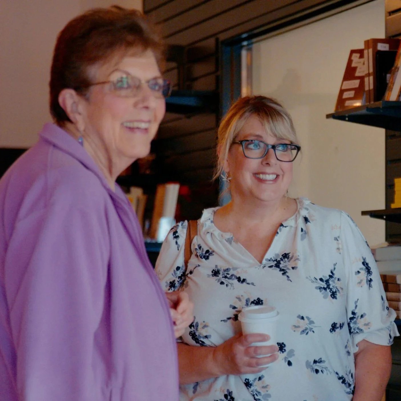 Two women smiling and talking, one holding a coffee cup, in a library or bookstore setting.