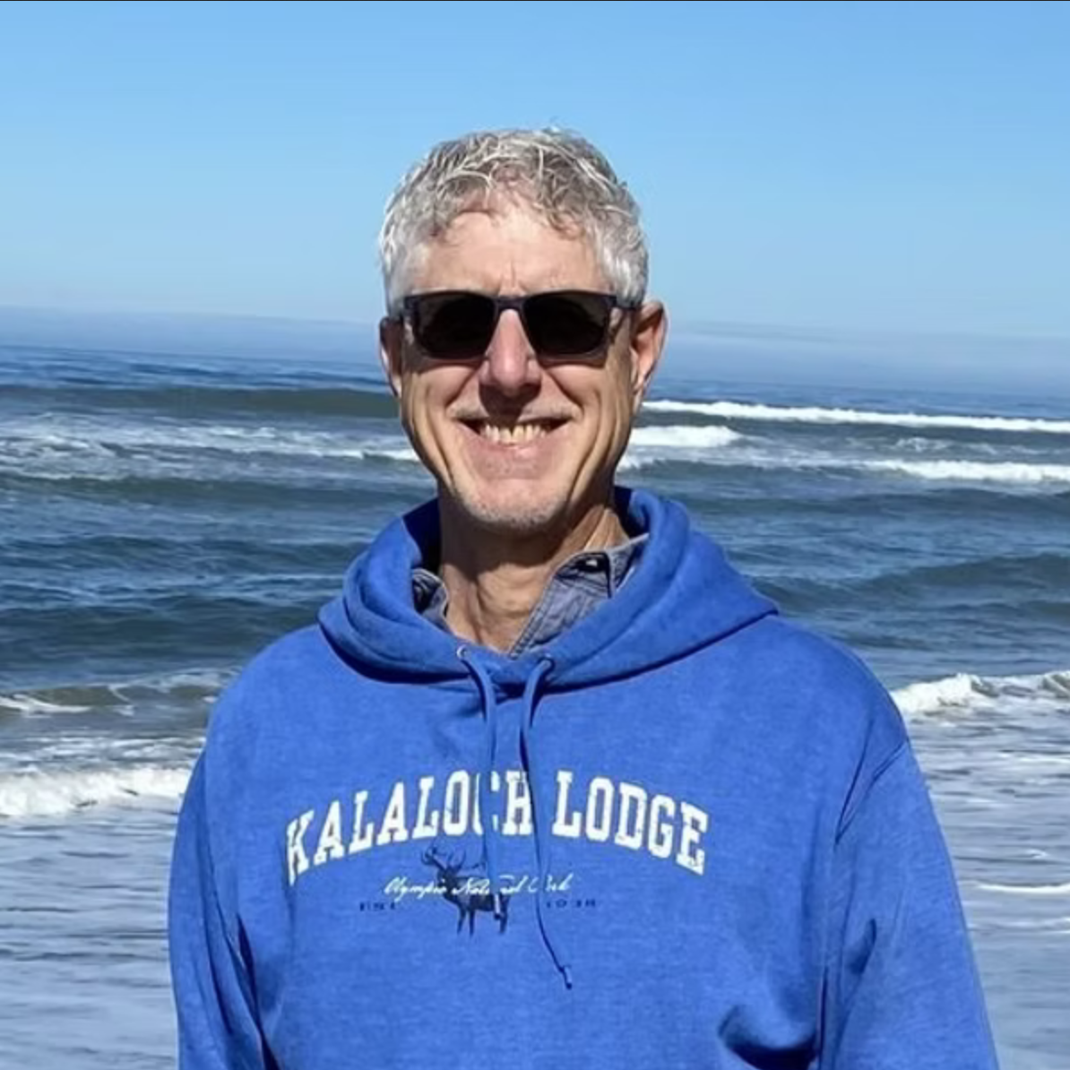 Paul Botsford: a man smiling at the beach, wearing sunglasses and a blue hooded sweatshirt, ocean waves in the background.