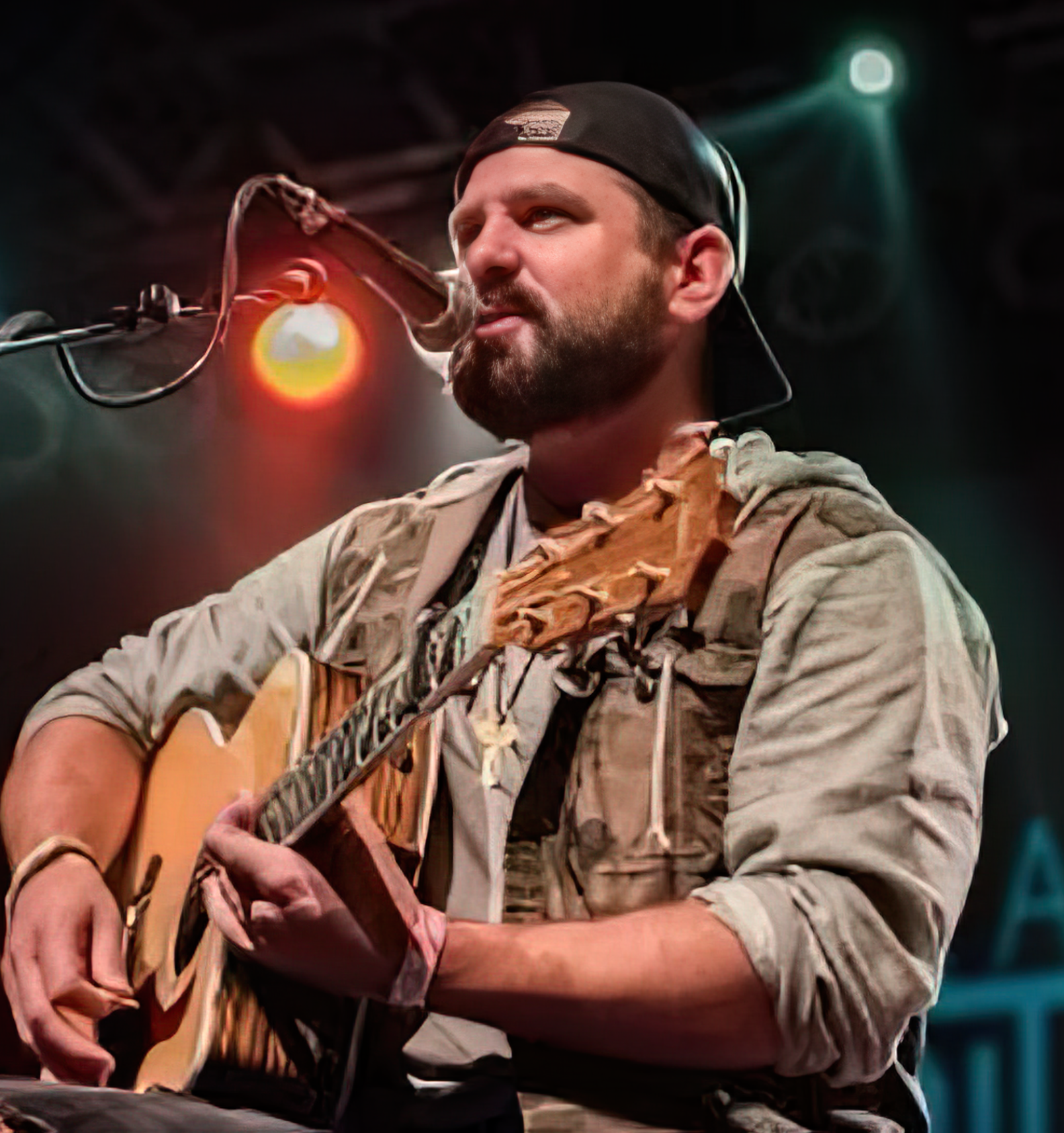 A man with a beard and wearing a black cap playing an acoustic guitar on stage, illuminated by stage lights.