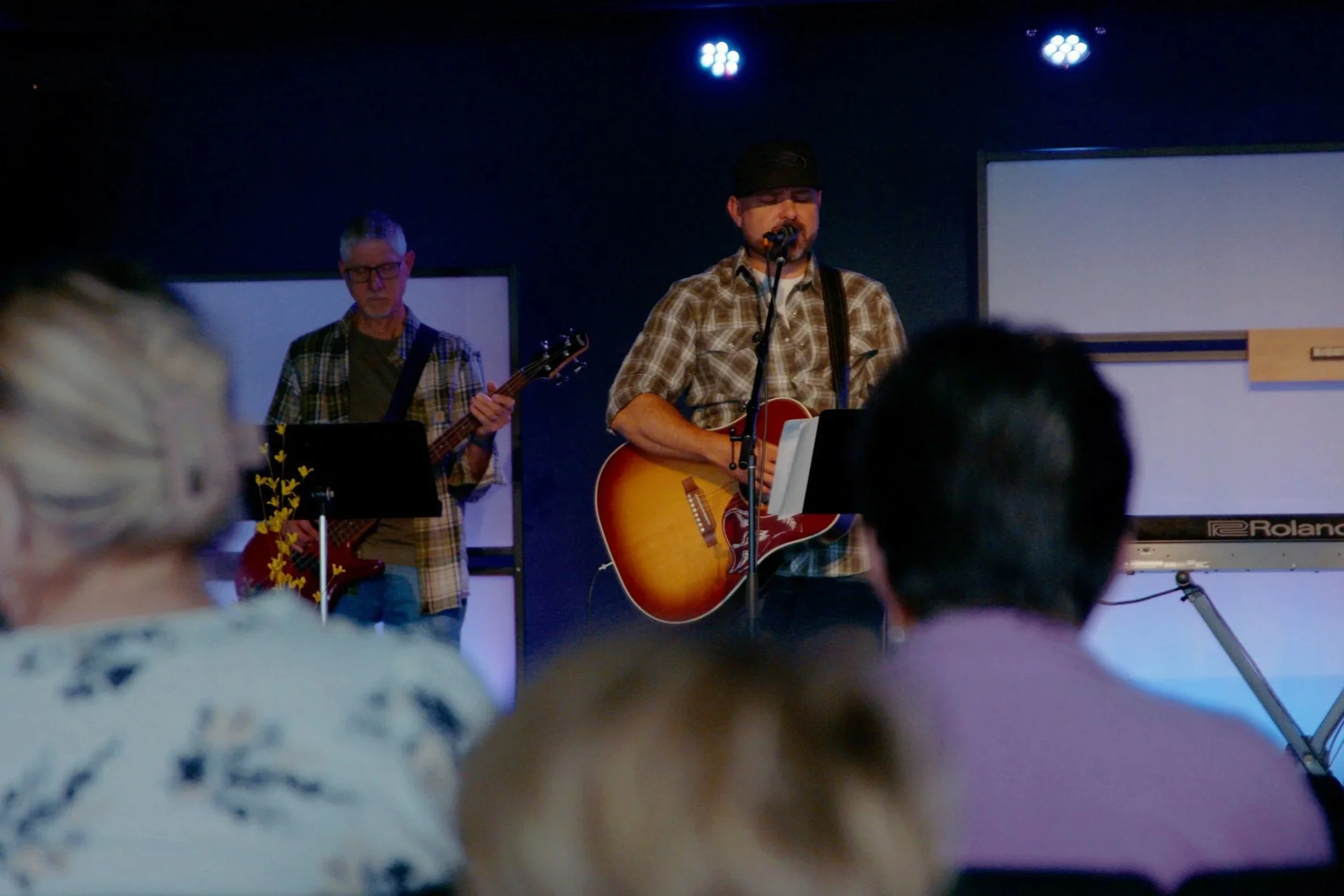 A band performing live on stage: a man singing and playing an acoustic guitar, and another man playing a bass guitar, with stage lights overhead and audience members in foreground.