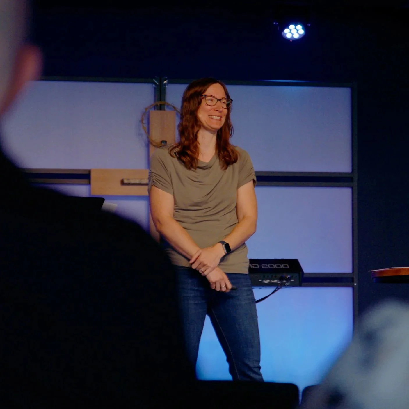 Woman with long brown hair and glasses smiling on stage during a sermon.