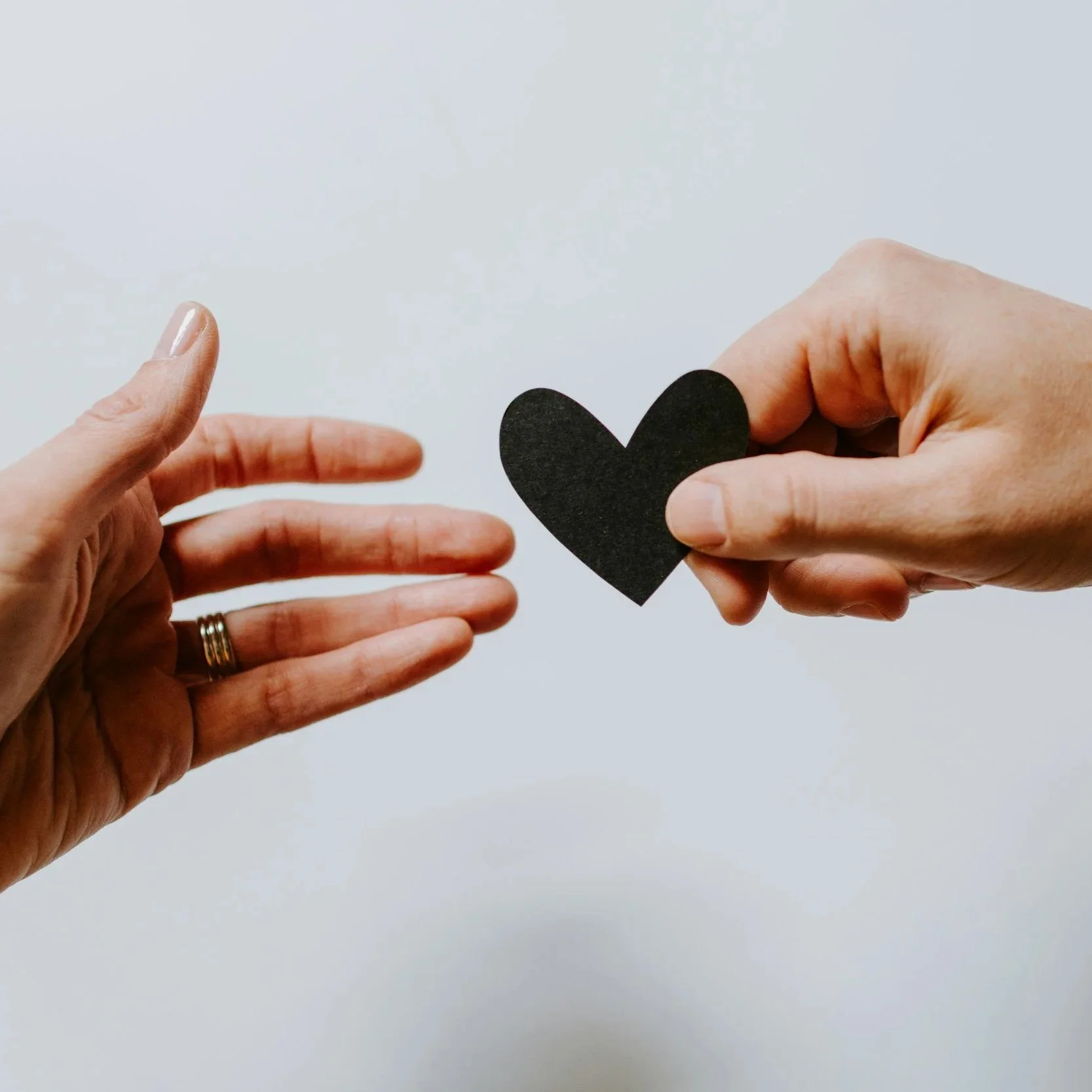 Two hands reaching towards each other, one holding a black paper heart, against a plain light background.