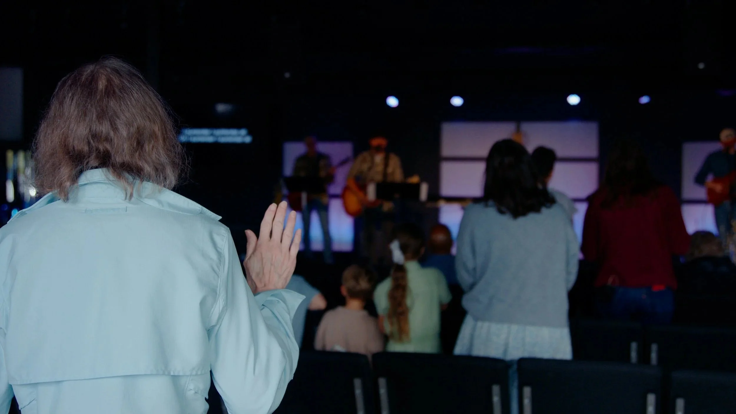 A woman with shoulder-length brown hair is standing in front of a group of people in a dark room with a stage. She appears to be praying or raising her hands in worship. The audience is seated, and there are musicians or speakers on stage.