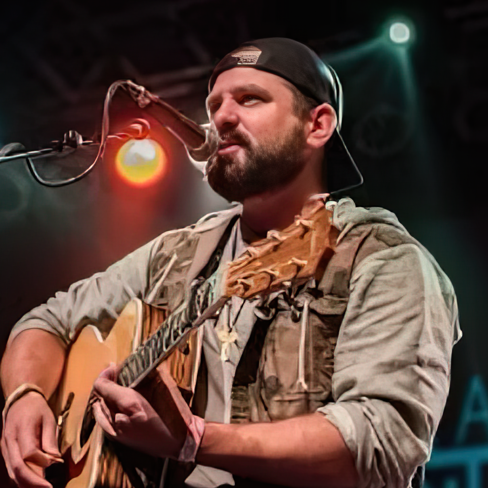 Nathan Botsford, a man with a beard and wearing a backwards black cap plays an acoustic guitar on stage, with colorful stage lights in the background.
