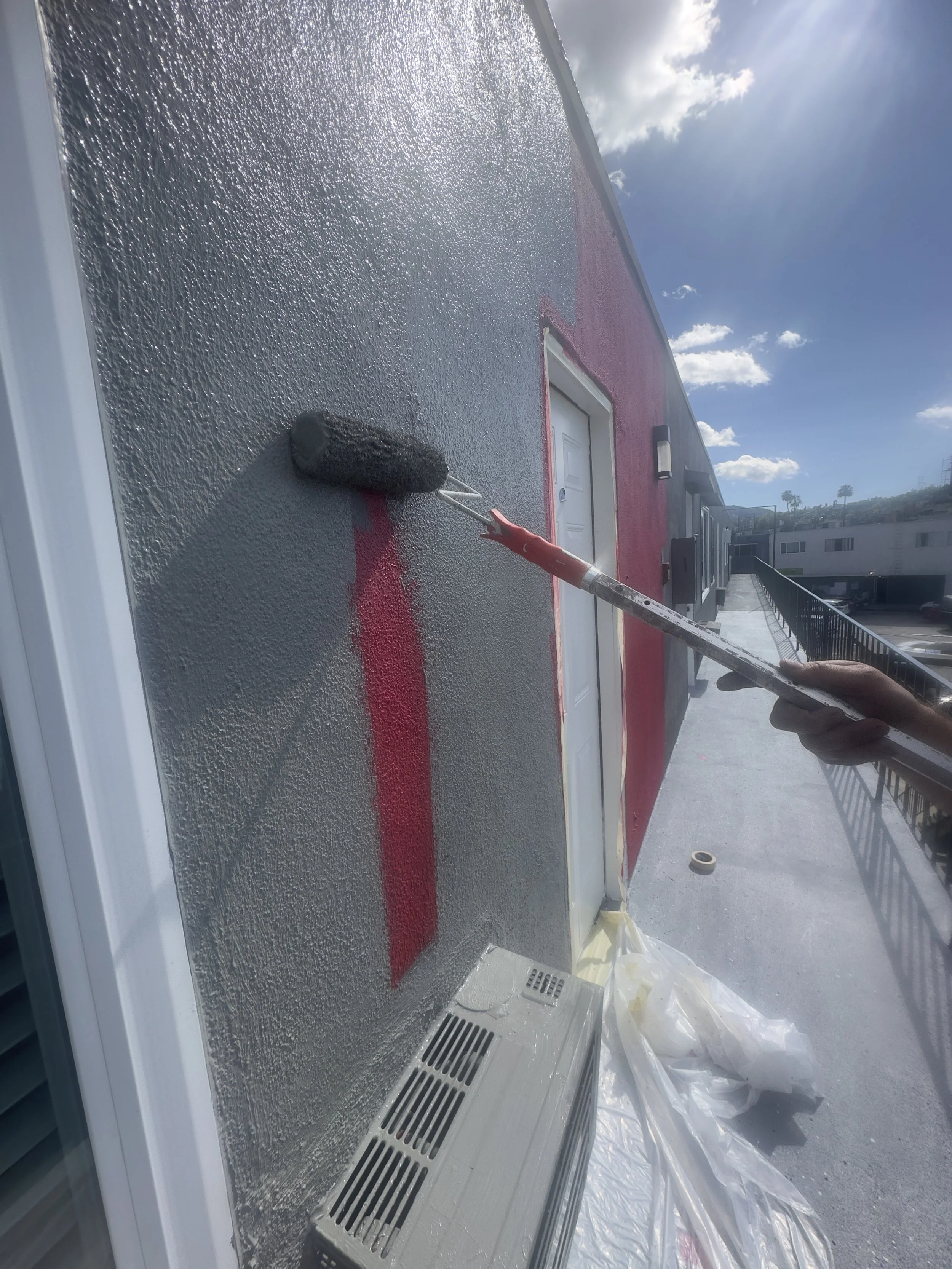 Persona pintando una línea roja en una pared exterior con rodillo en un espacio con cielo nublado.