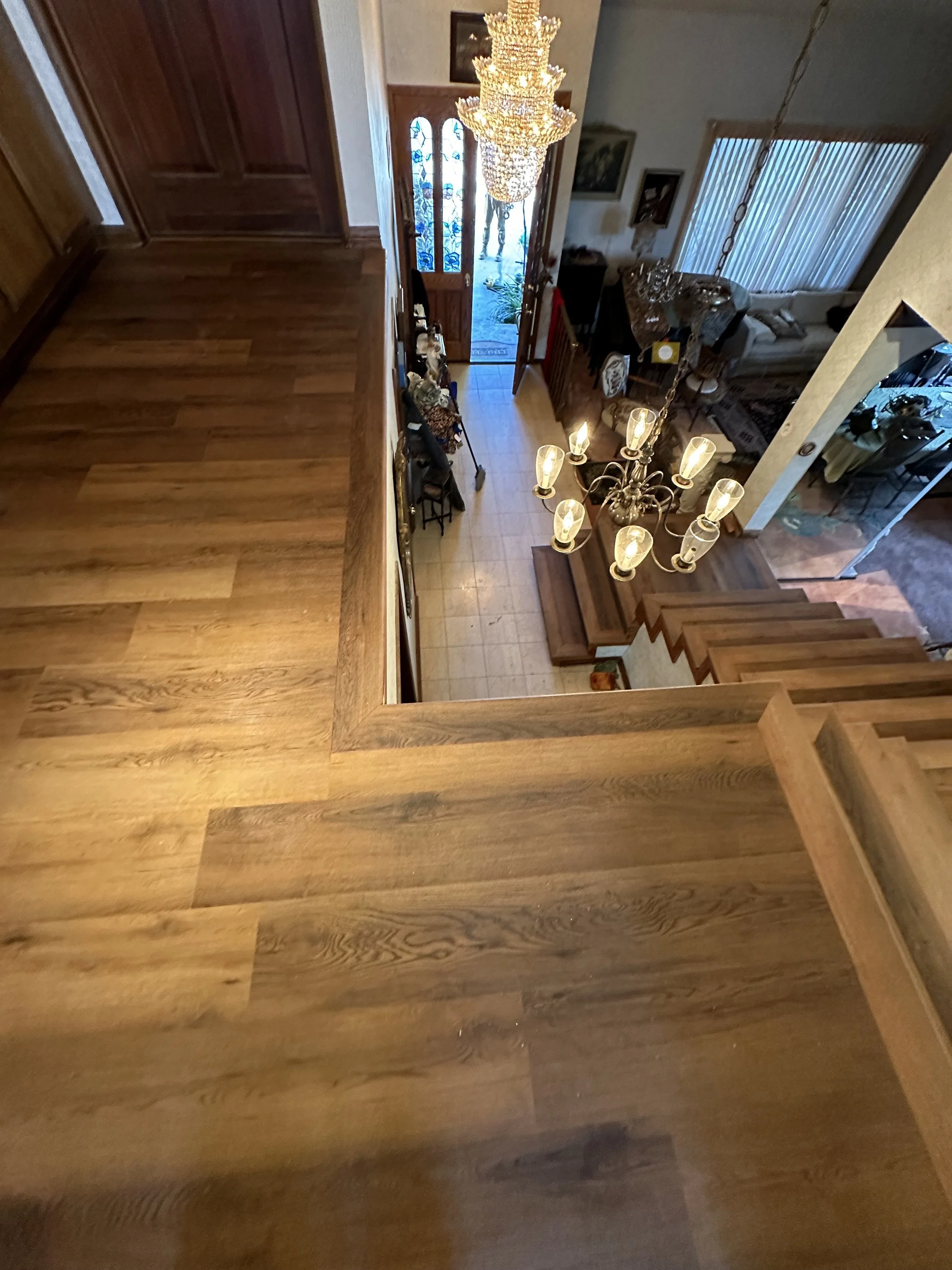 Vista desde arriba de un interior de casa con pisos de madera, una lámpara grande de cristal, una entrada con puerta de madera y ventanas con vitrales, y una sala con muebles.