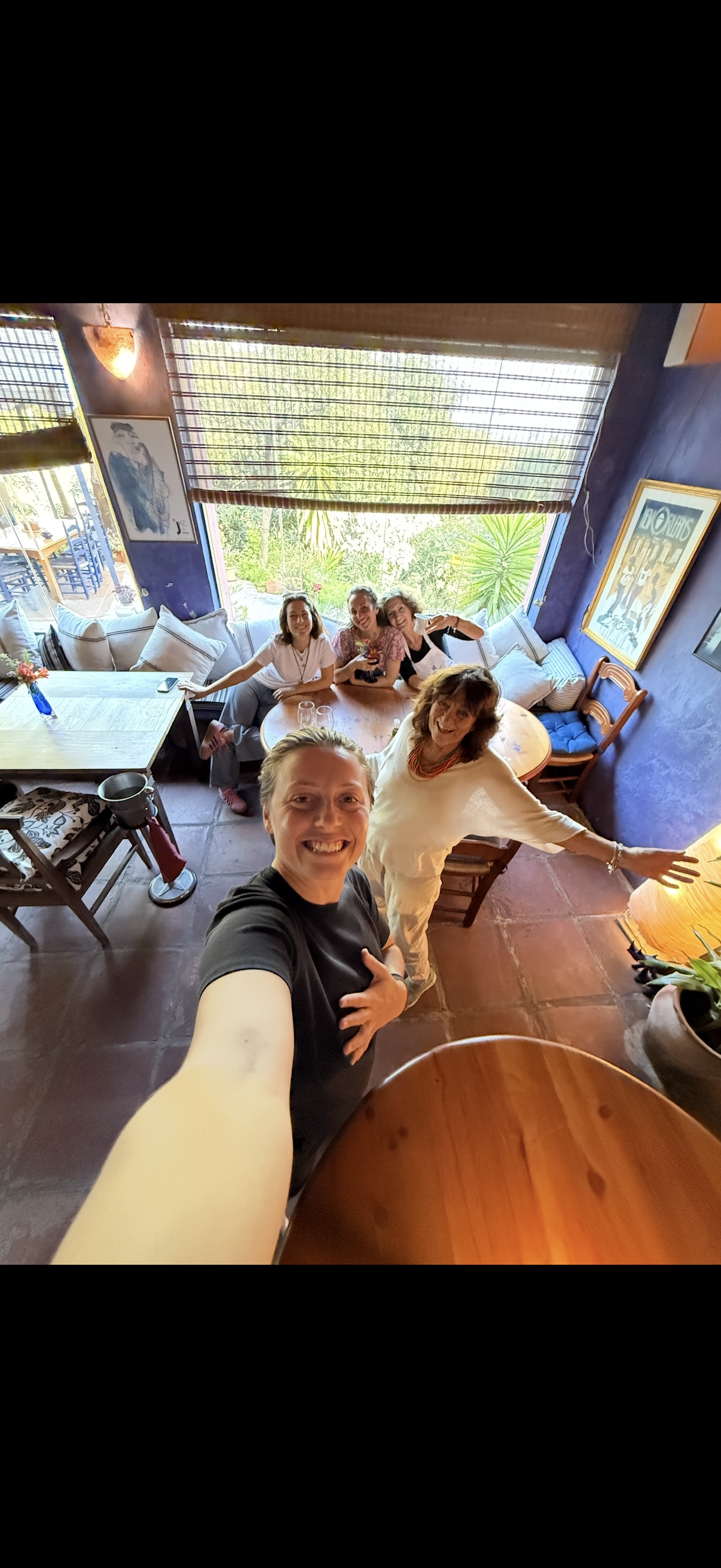 Group of five women smiling and taking a selfie in a cozy restaurant with purple walls, large windows, and tropical plants outside.