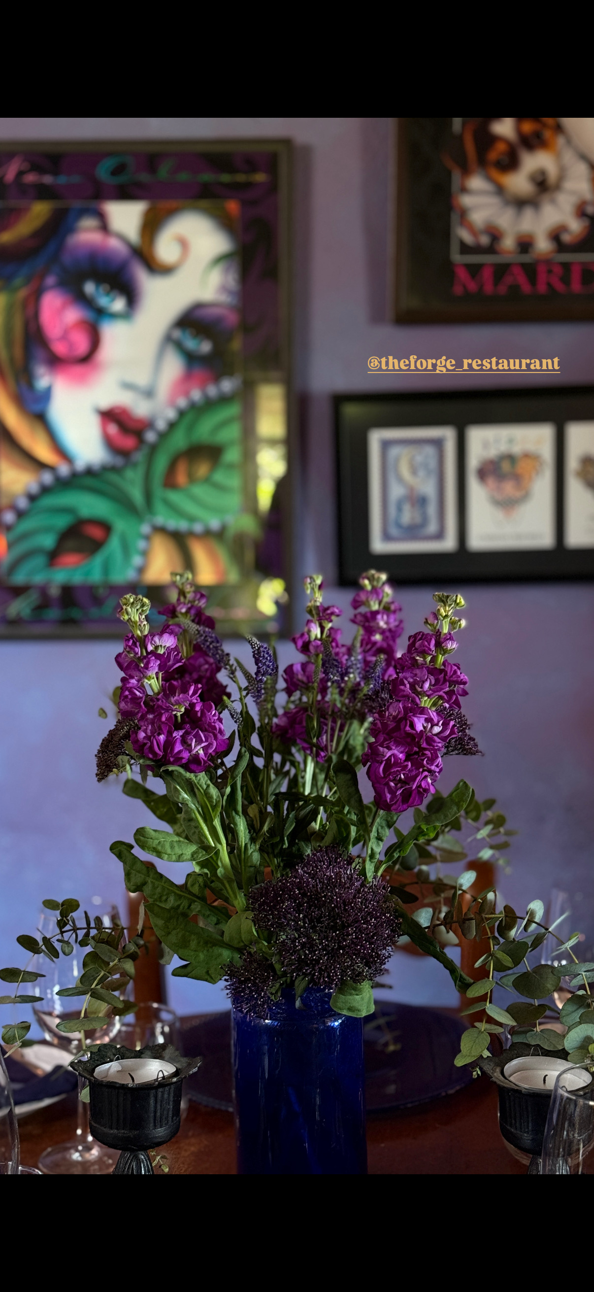 A vase with purple flowers and greenery on a table in front of framed artwork and colorful paintings hanging on a purple wall.