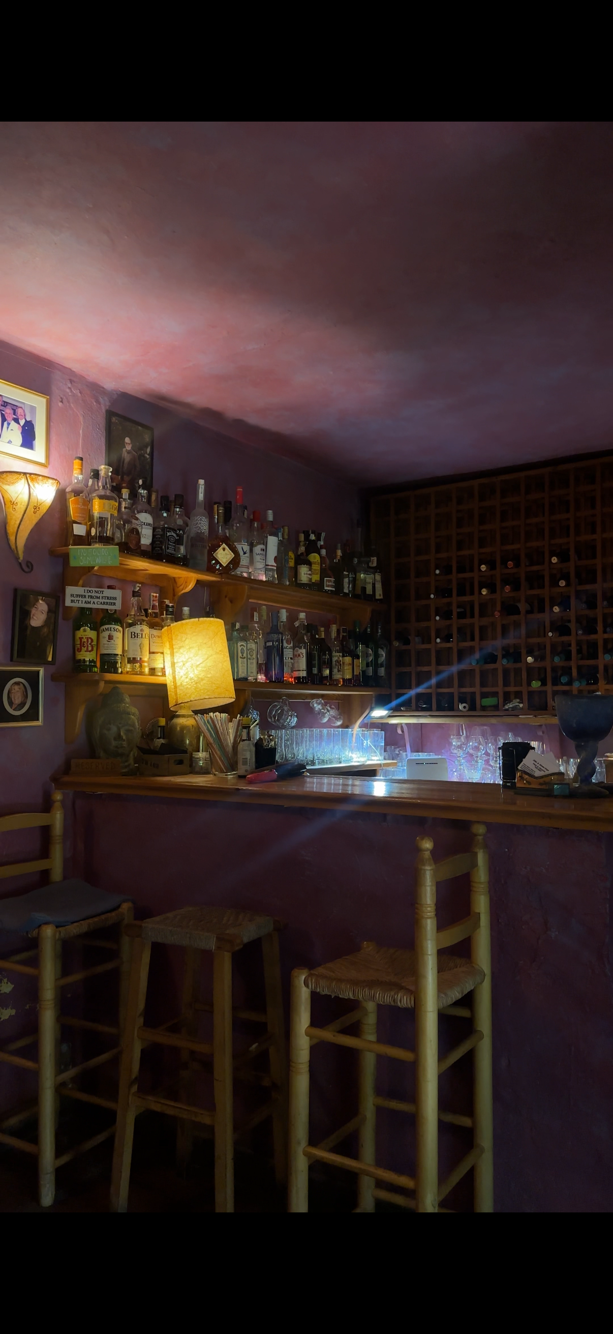 Interior of a cozy bar with a wooden counter, barstools, shelves filled with various liquor bottles, wine racks, a Buddha statue, framed photos, and warm lighting.