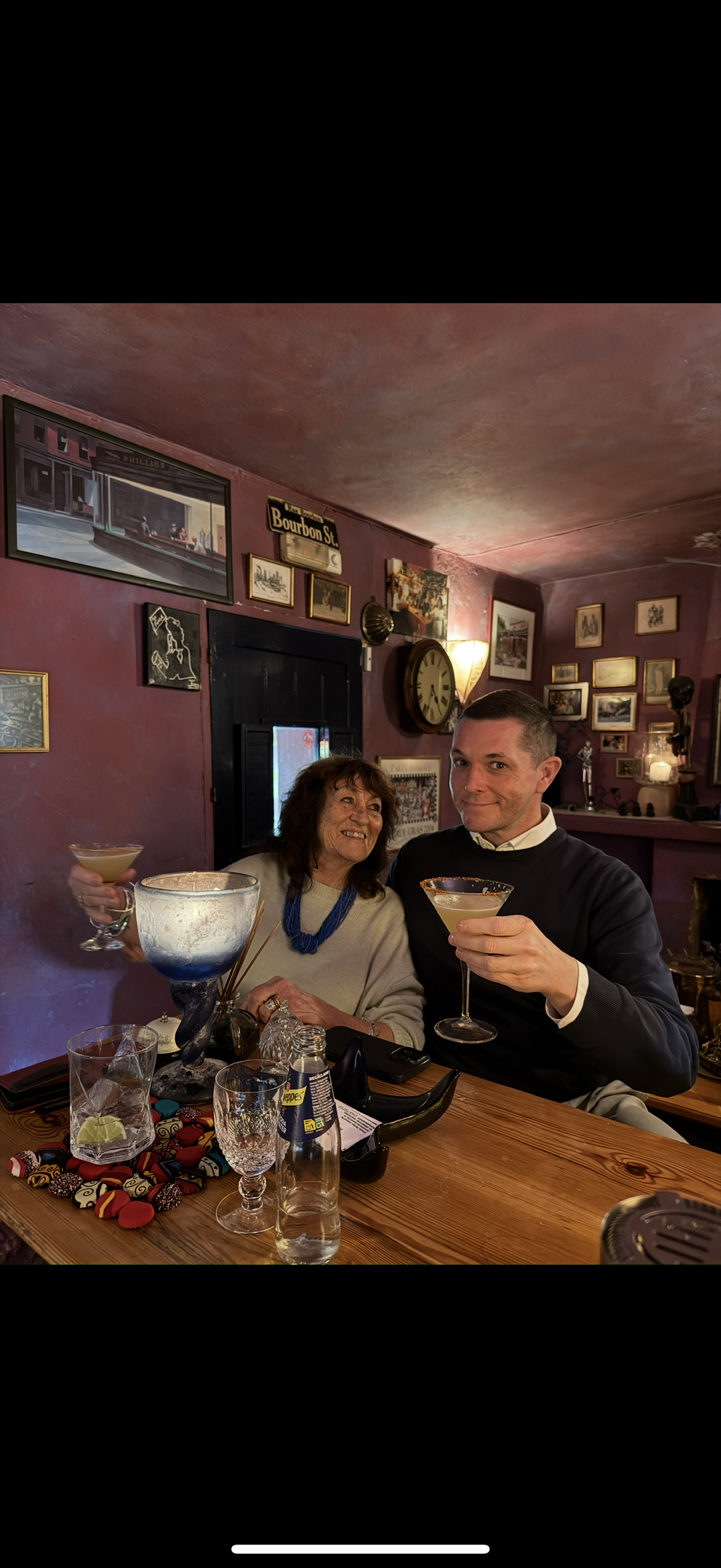 Two people sitting at a table holding cocktails inside a decorated room with framed pictures and a large clock on the wall.